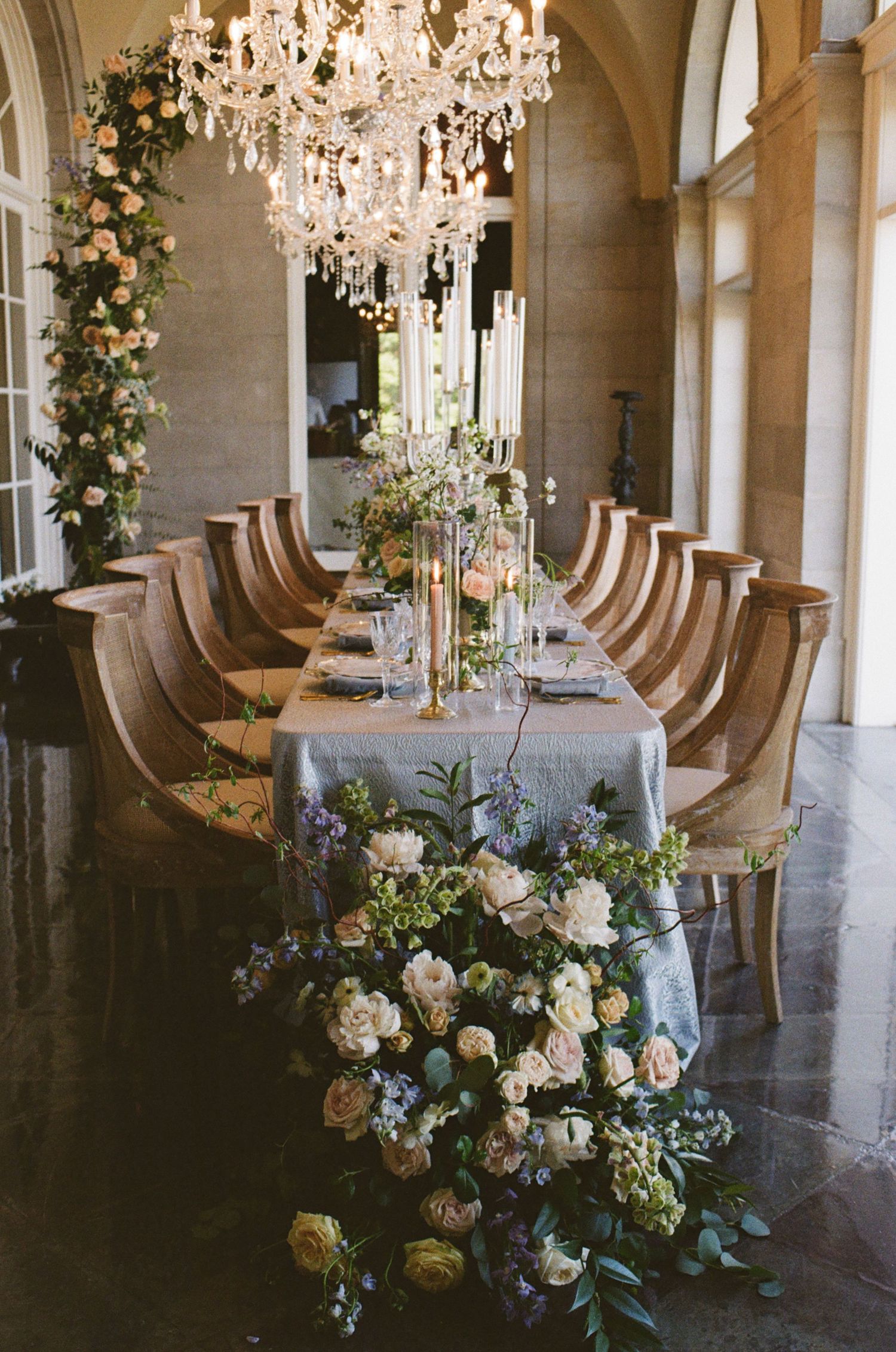 Elegant wedding reception table with cascading floral arrangements, crystal chandelier, and French-style chairs in a grand venue.