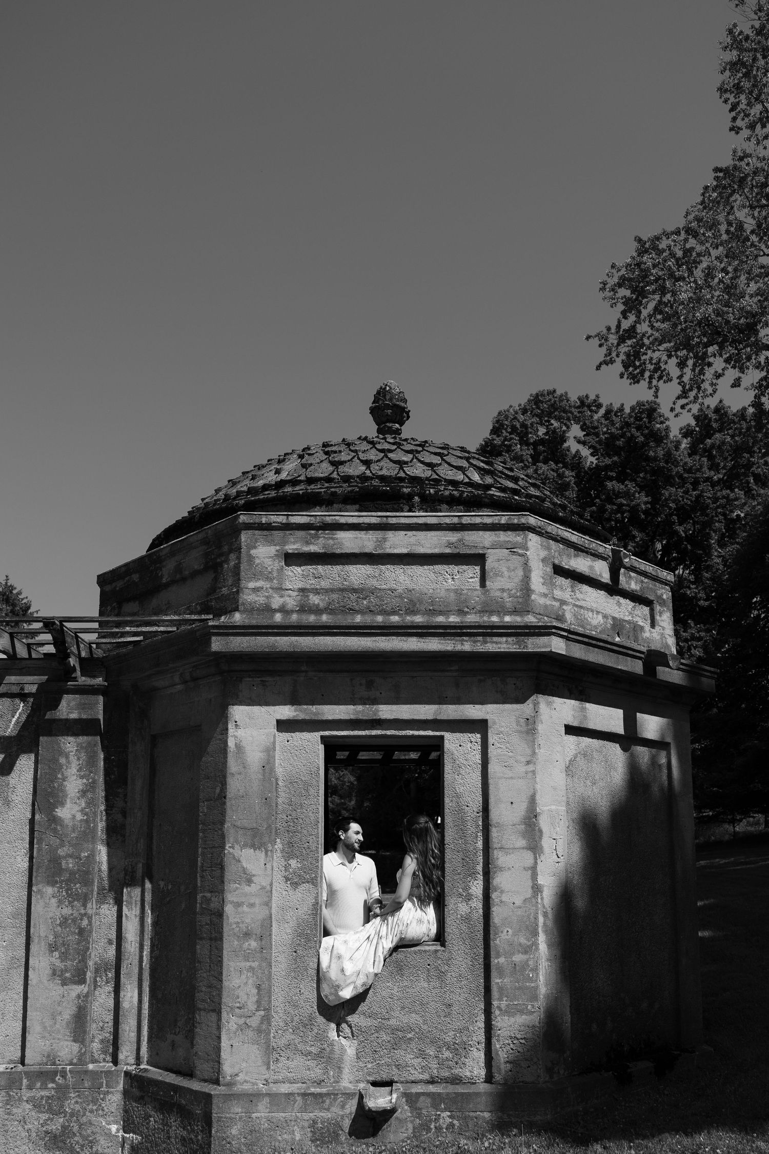 Black and white photo of a couple embracing at the doorway of an ornate stone gazebo surrounded by trees.
