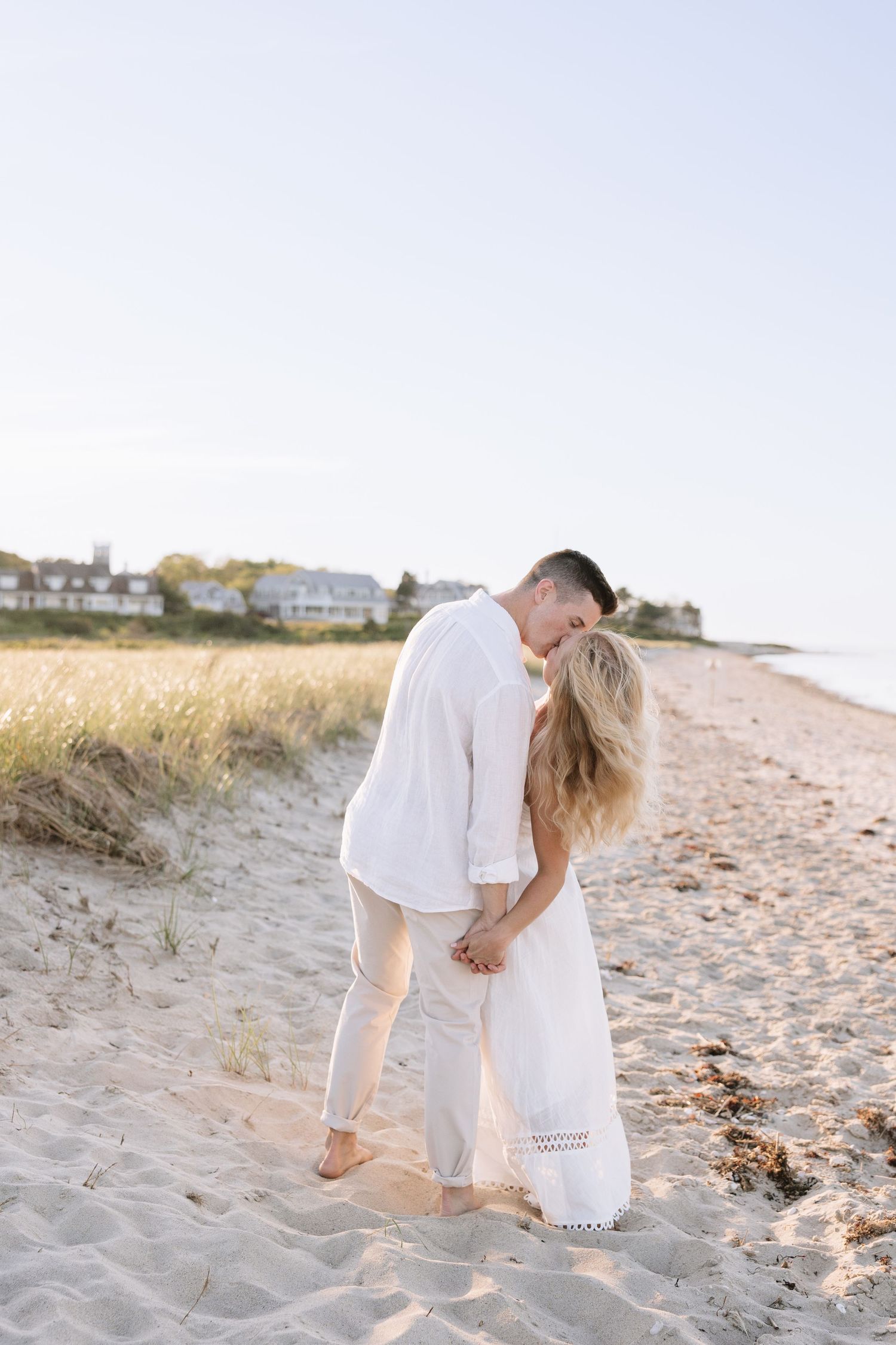 Couple in white outfits sharing a romantic kiss on a sandy beach at golden hour with coastal homes in the background.