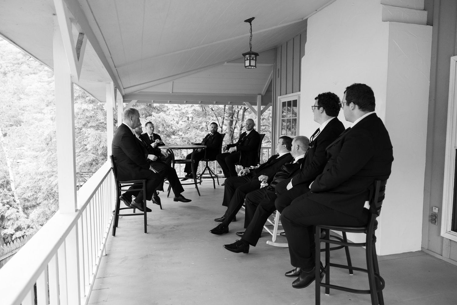 Groomsmen in black suits relaxing on a porch before a wedding ceremony in a black and white photo.