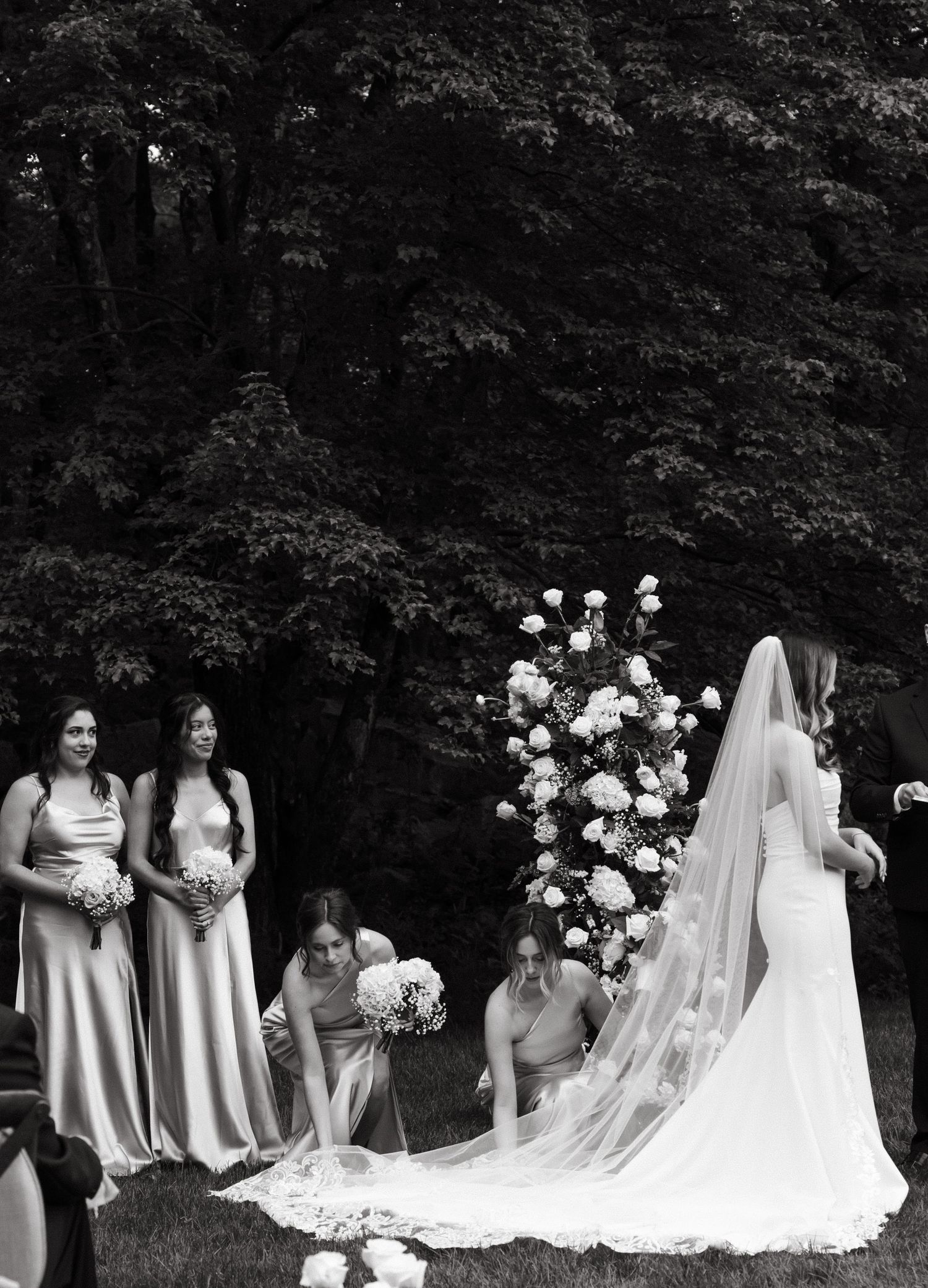 Wedding party poses outdoors with bridesmaids in light dresses and a bride in white gown against wooded backdrop.