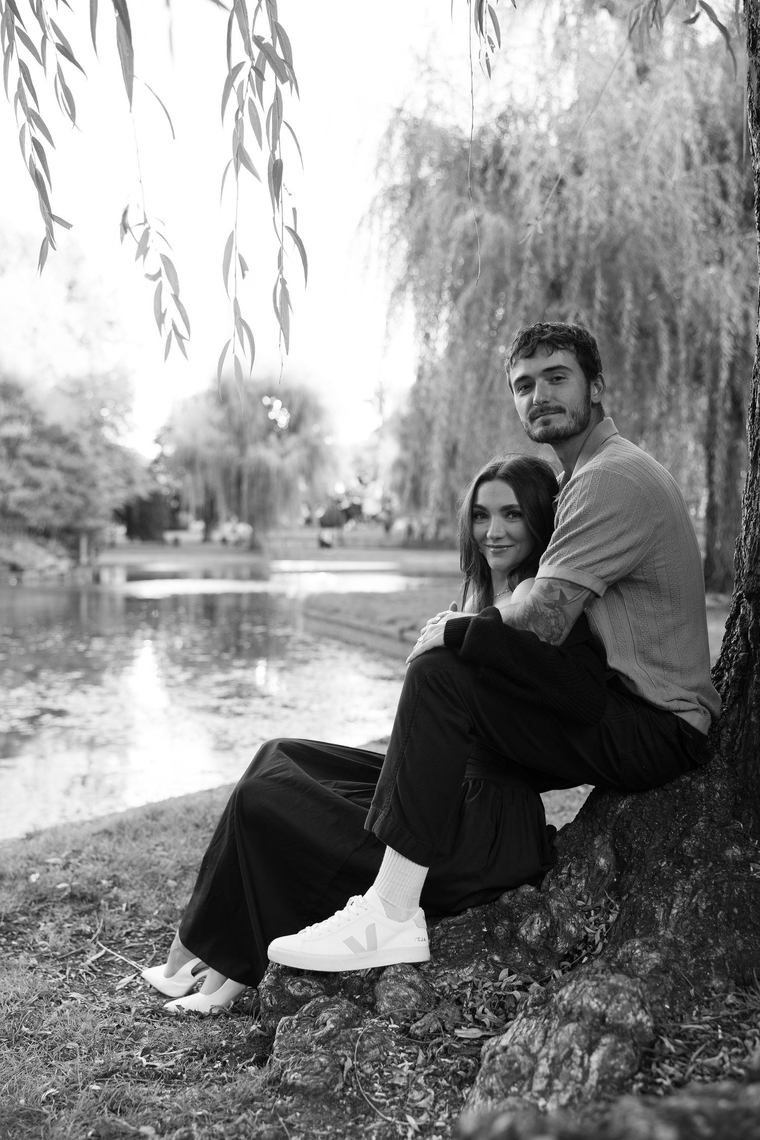 Two people relax against a tree trunk by a pond with willow branches hanging above.