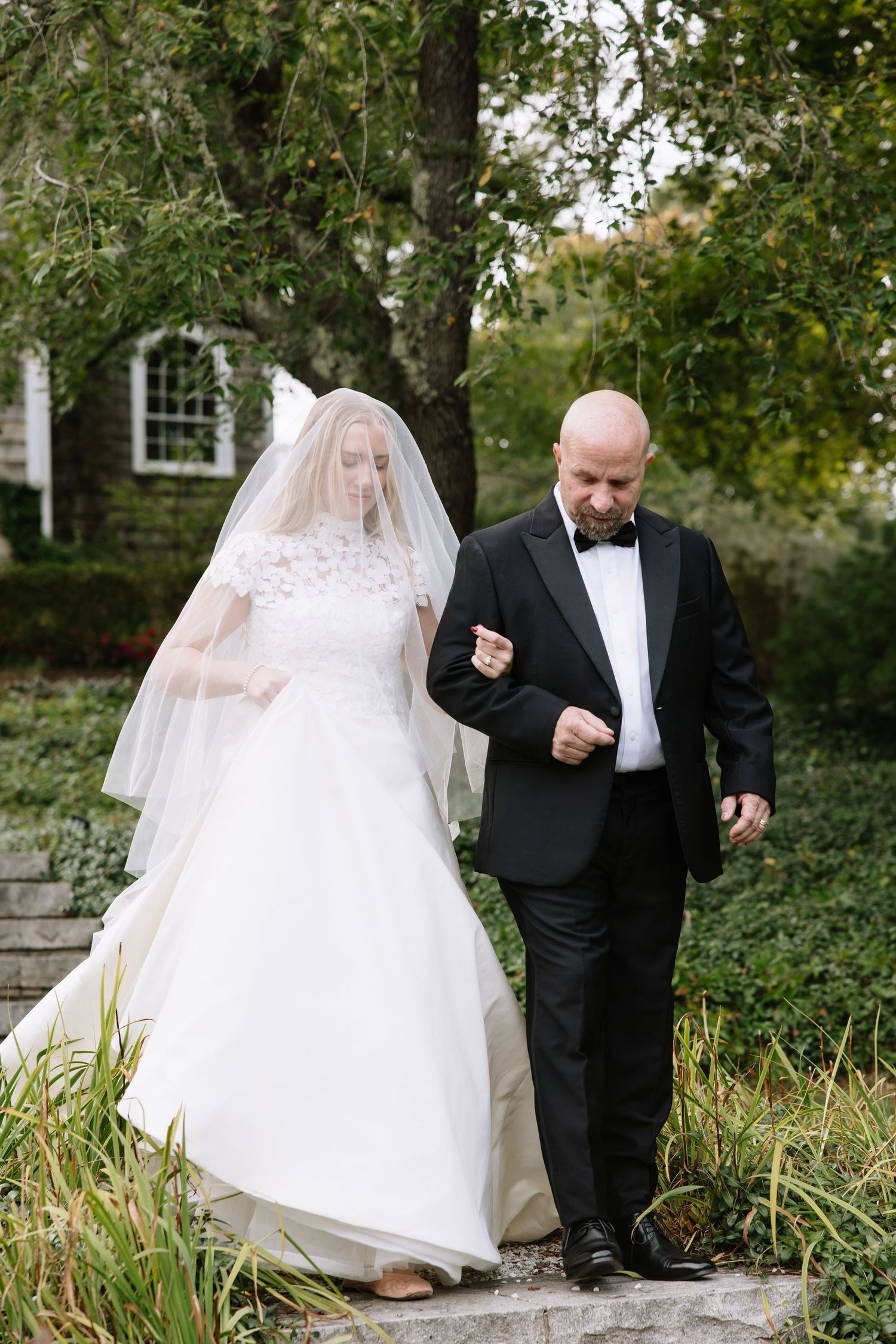 Bride in lace gown walks down stone steps with escort at outdoor wedding venue.