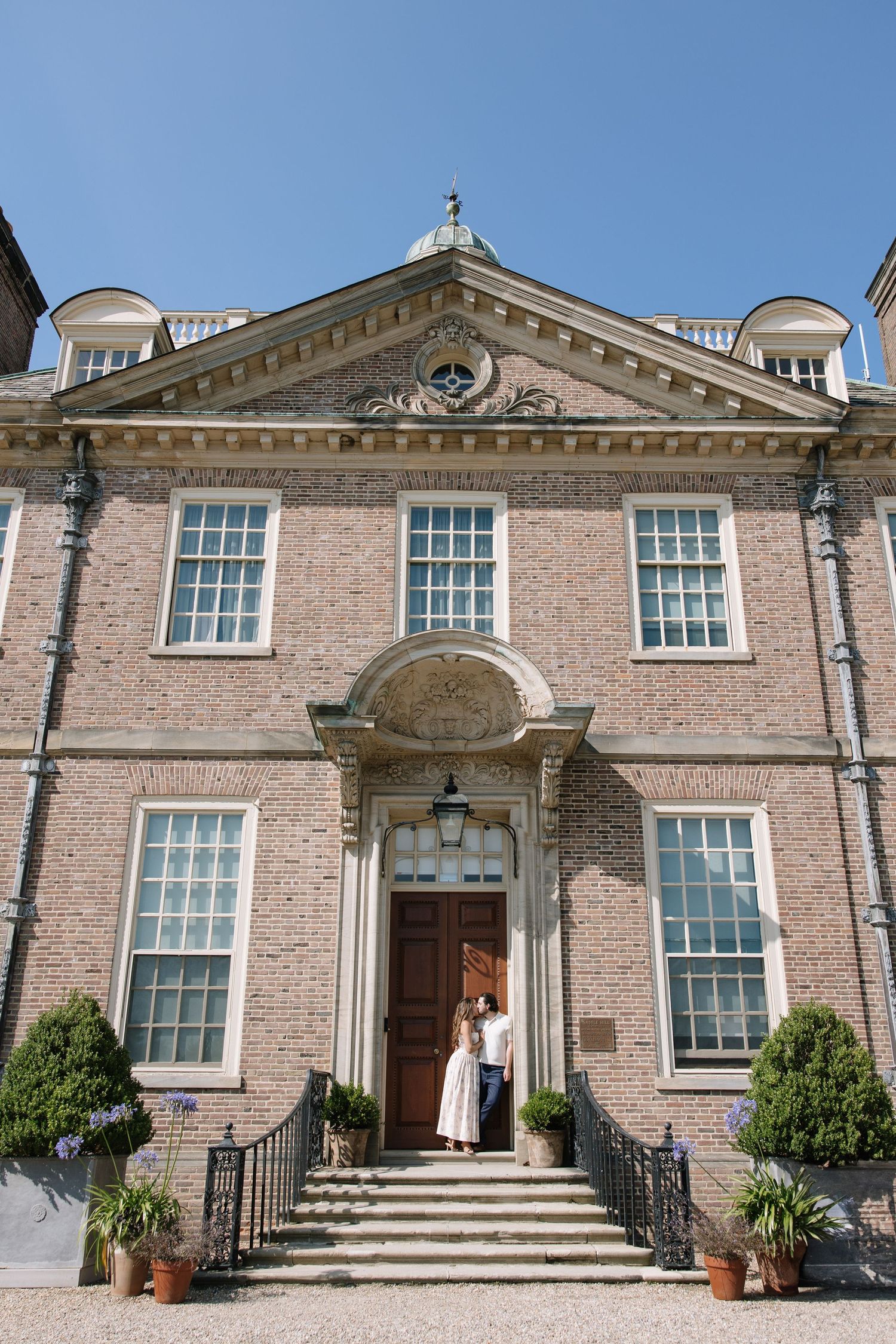 Couple standing at entrance of elegant Georgian brick manor house with pediment and sash windows under blue sky.