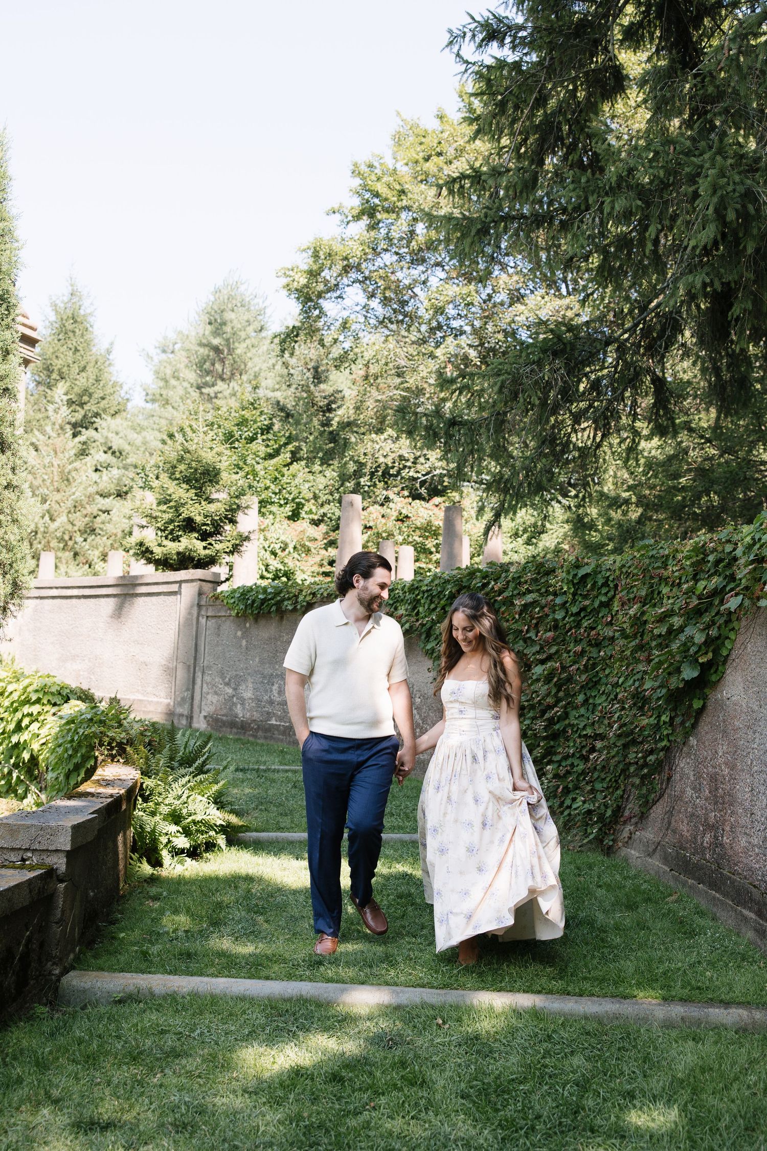 A couple holds hands walking through a lush garden, woman in floral maxi dress, man in white polo and navy pants.