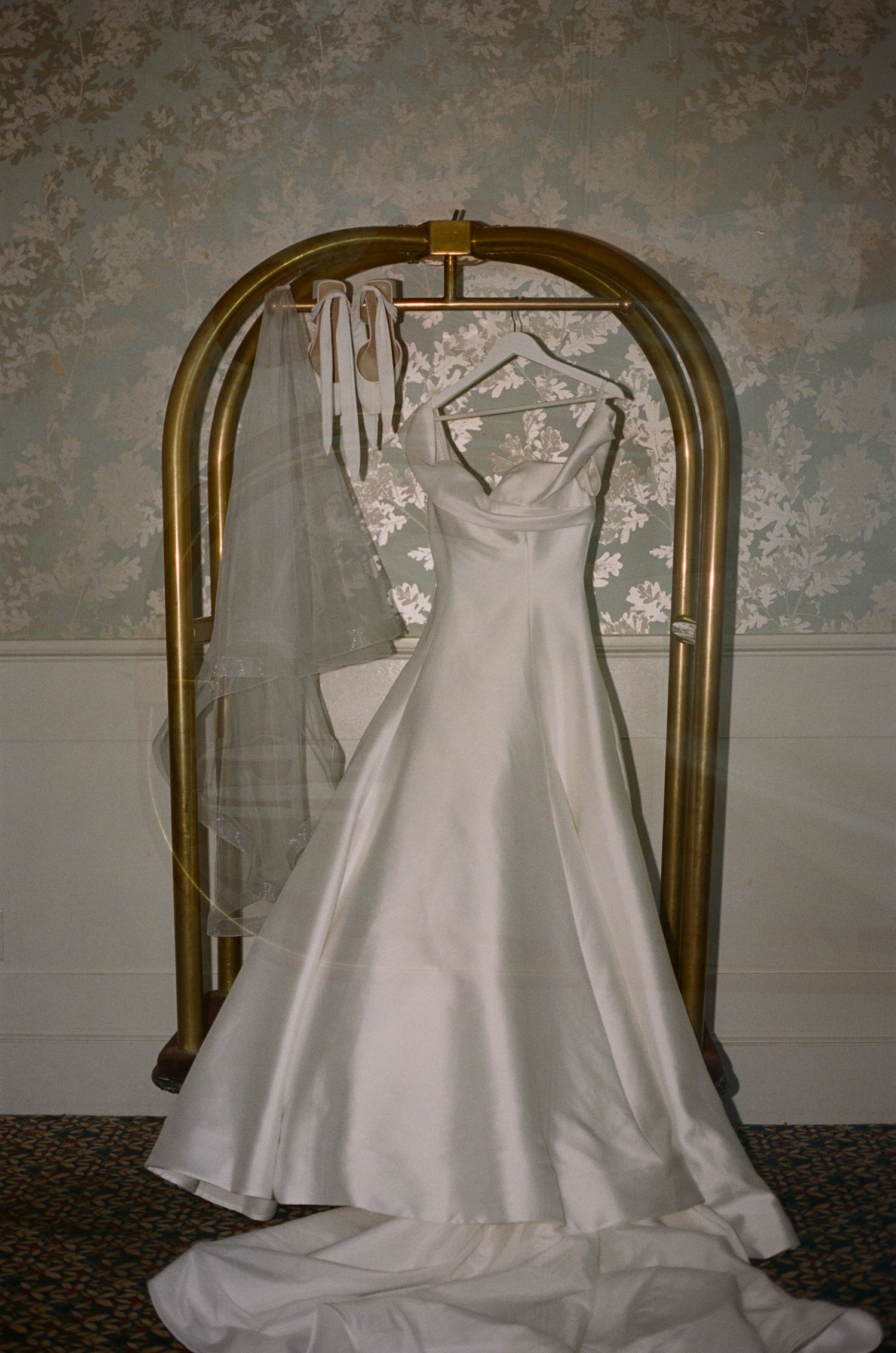Elegant white satin A-line wedding dress with train hanging on gold luggage rack with veil in hotel room.