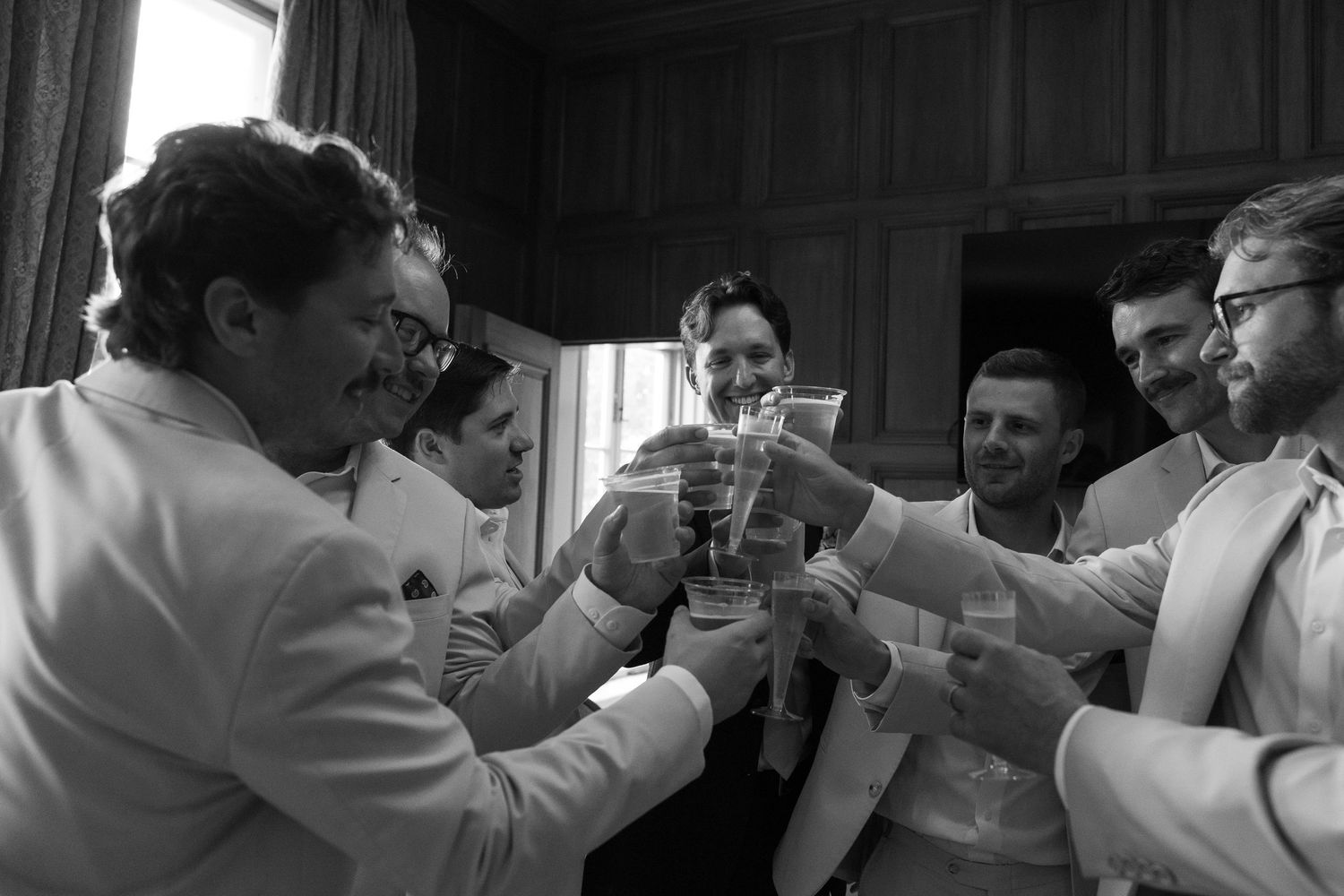 Groomsmen in white suits toast with drinks in a black and white wedding celebration photo.