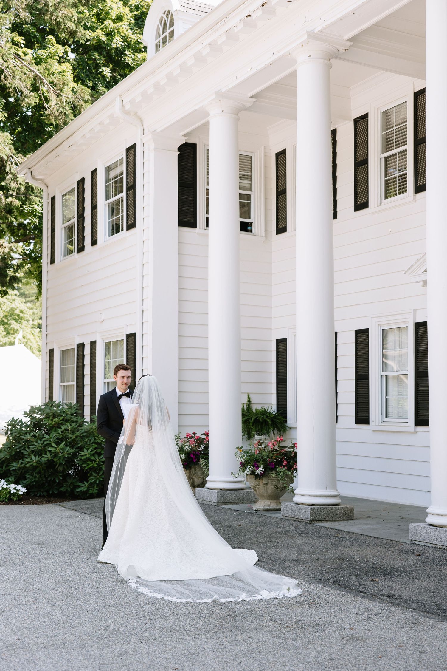 Bride and groom pose in front of elegant white colonial mansion with tall columns and black shutters on their wedding day.
