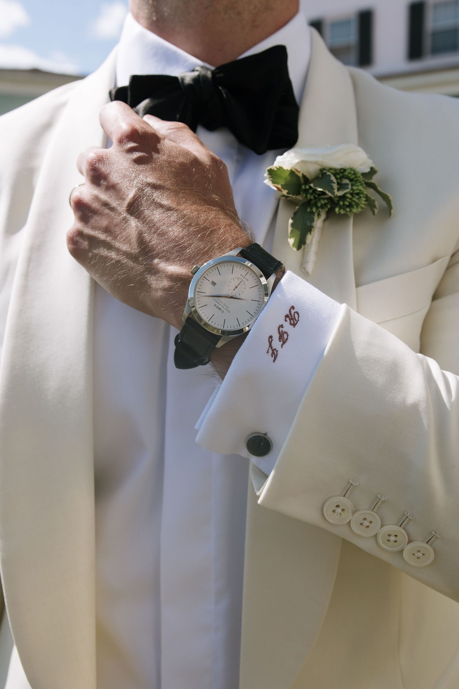 Groom in white tuxedo jacket adjusting black bow tie, wearing monogrammed cufflinks, watch, and white boutonniere.