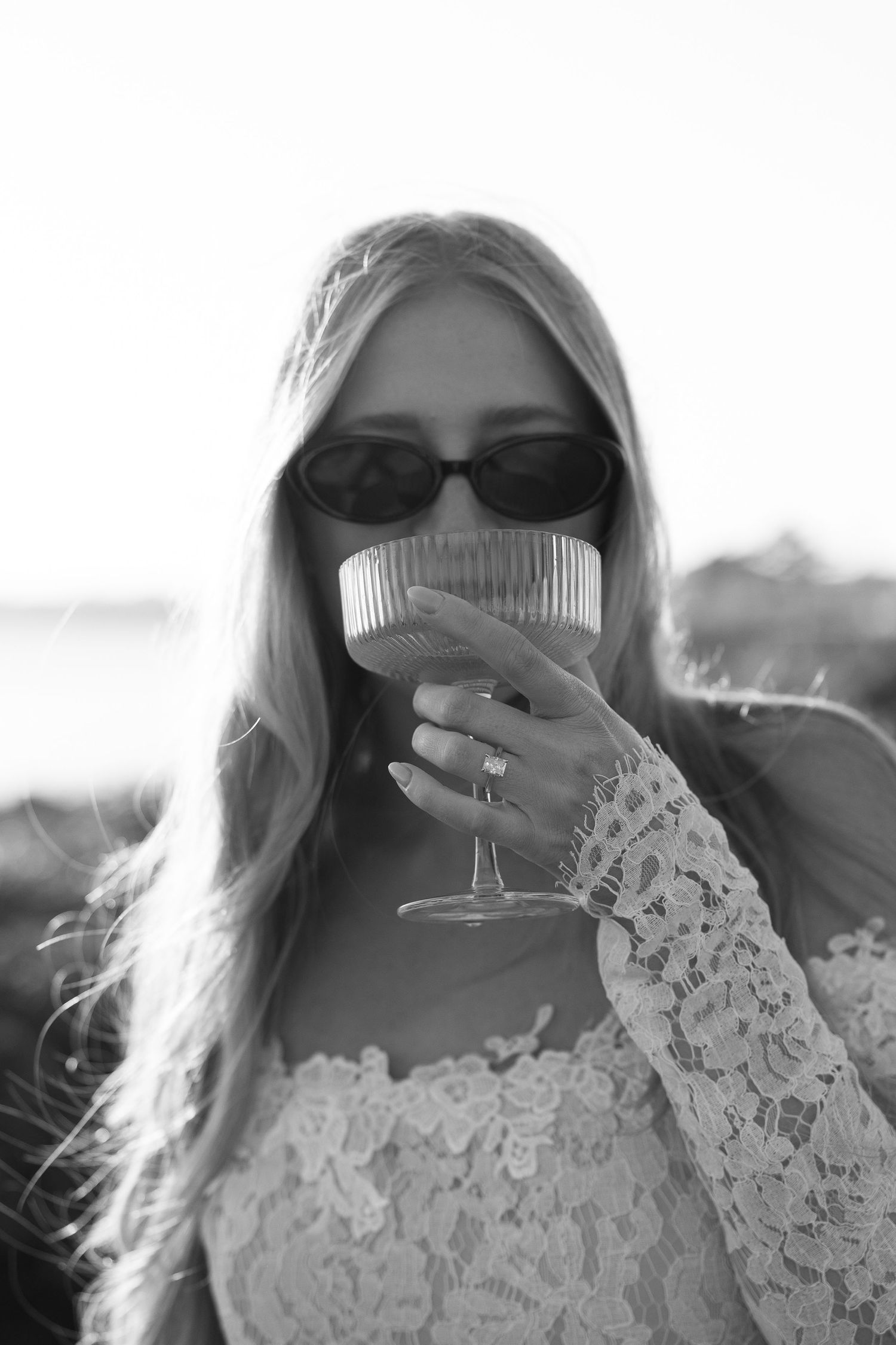 Blonde woman in lace dress and sunglasses sipping from elegant coupe glass in black and white photo.