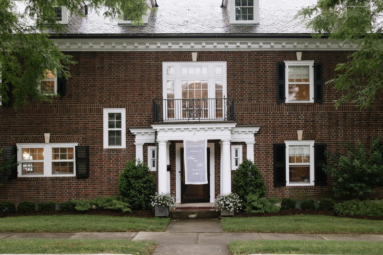 Elegant brick colonial home with white trim, black shutters, and manicured shrubs at the entrance.