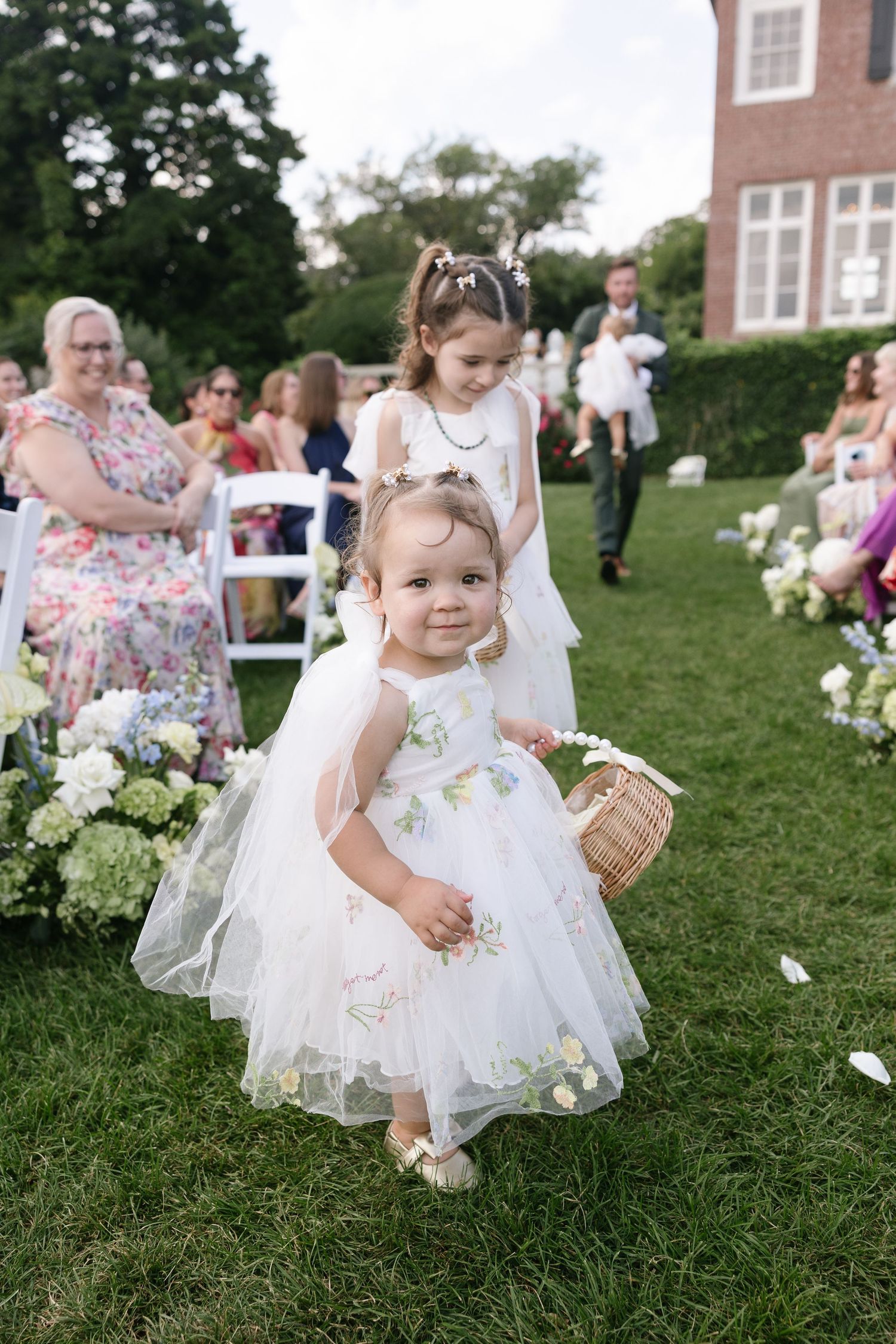 A toddler flower girl in a floral tulle dress carries a small wicker basket down a grassy wedding aisle past seated guests.