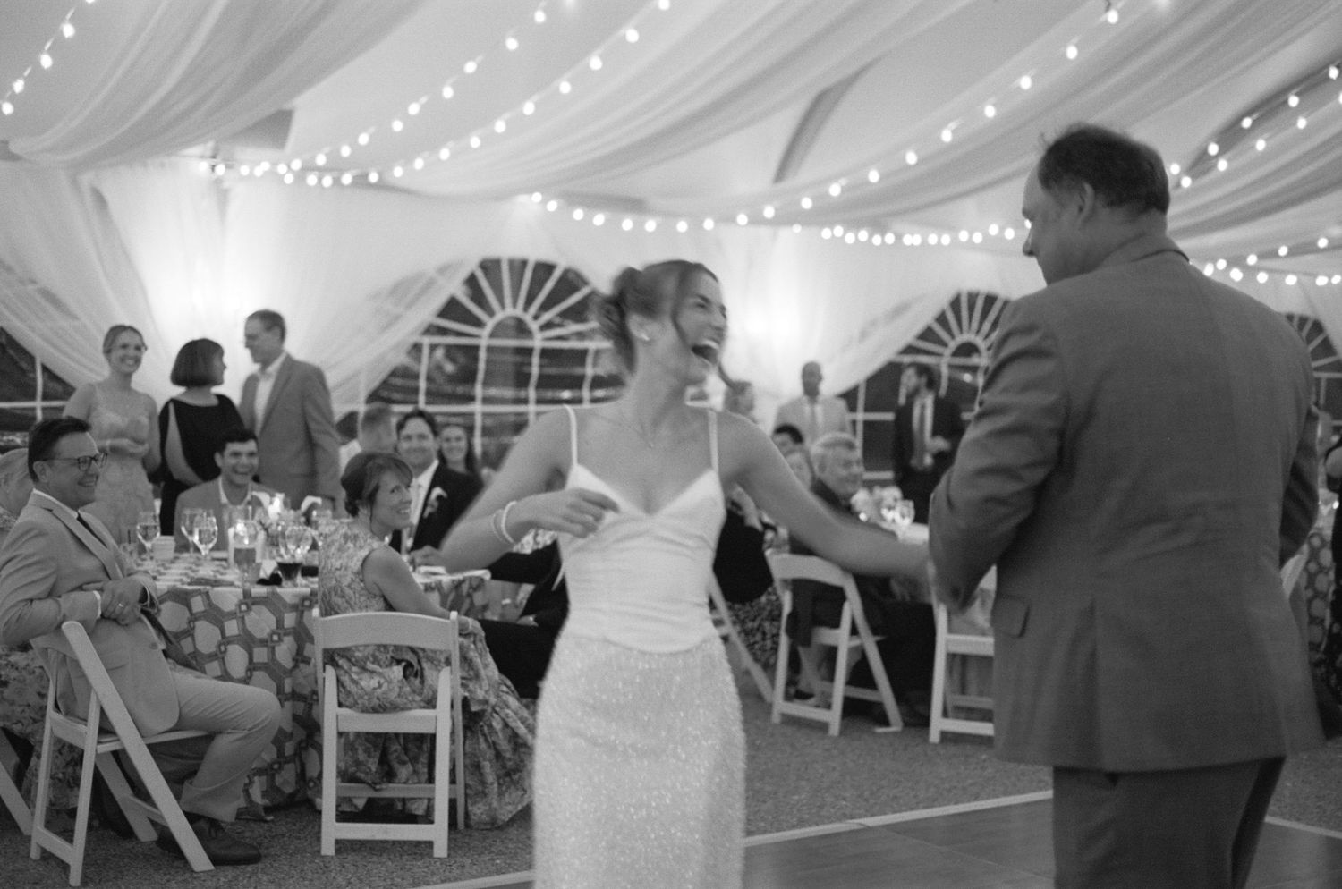Black and white photo of a bride in a gown dancing with a man in a suit at a tented wedding reception while guests watch from tables.