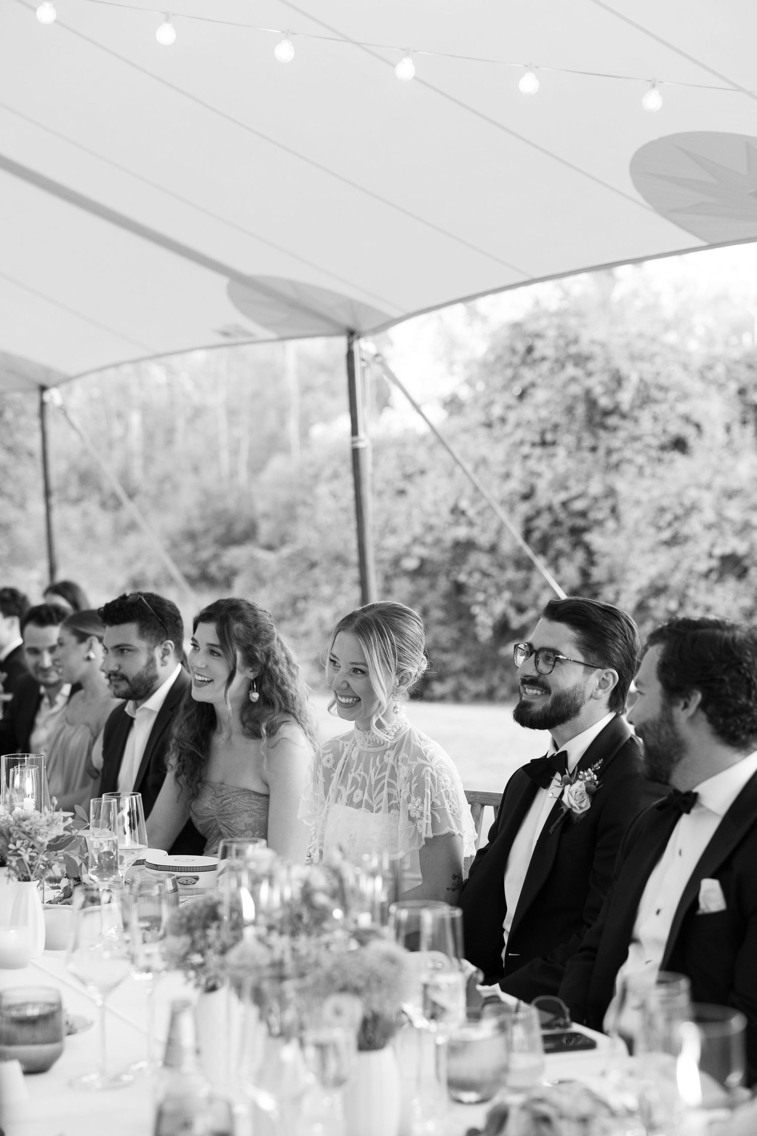 Black and white photo of wedding party seated at reception table under tent, laughing and enjoying celebration.