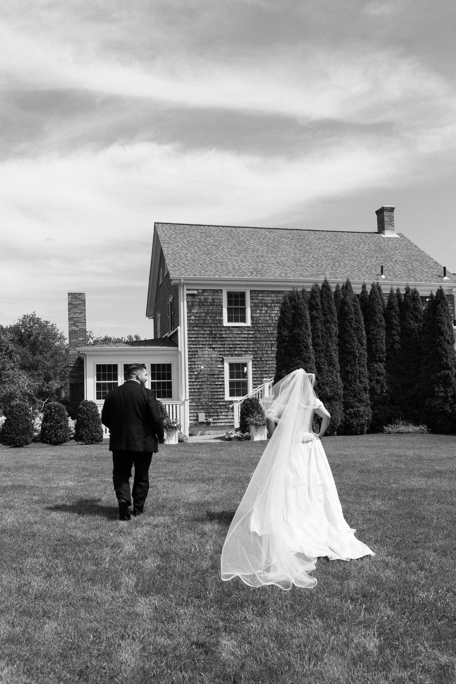 Black and white photo of bride and groom walking toward charming estate.