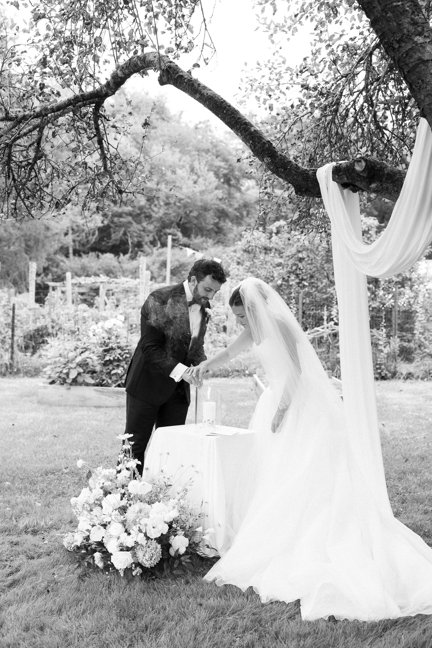 Black and white photo of bride and groom lighting a unity candle under a tree during outdoor wedding ceremony.