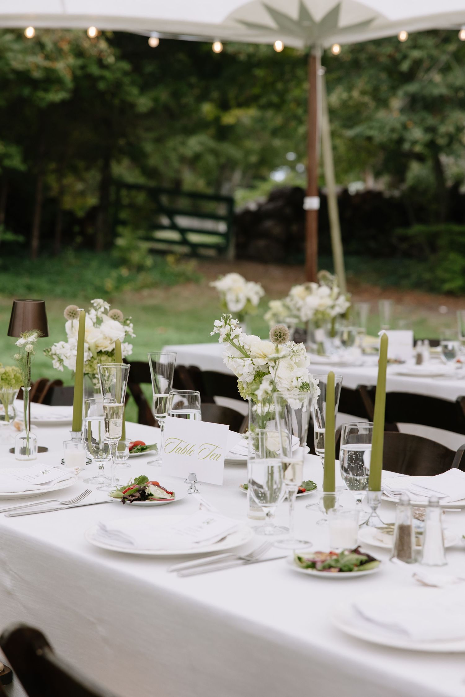 Elegant outdoor wedding reception table with white linens, green taper candles, white floral centerpieces, and glassware under a tent.