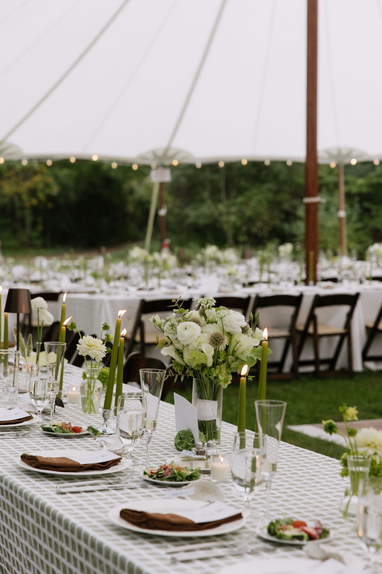 Elegant outdoor wedding reception under a white tent with green gingham tablecloths, white floral centerpieces, and string lights.