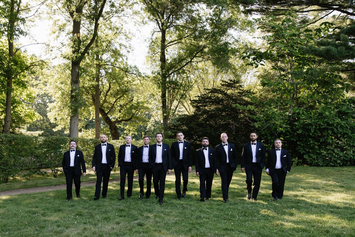 Groomsmen in black tuxedos lined up for portrait on sunny garden lawn.