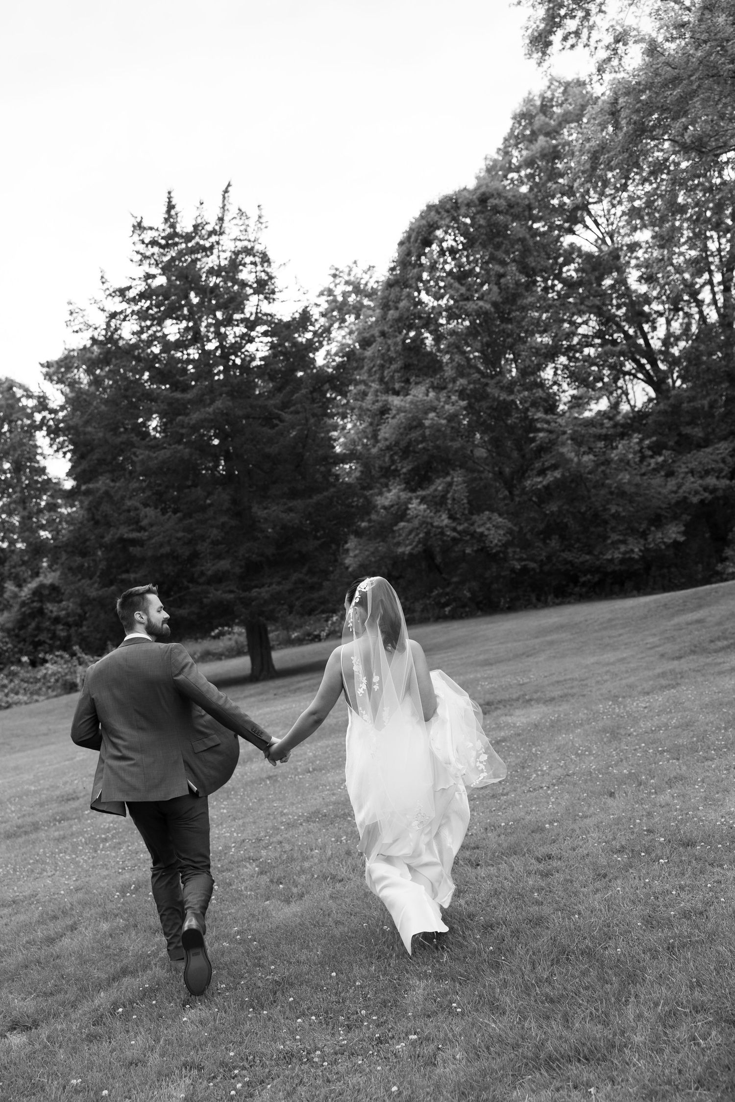 Black and white photo of bride and groom holding hands walking across a lush green lawn with trees.