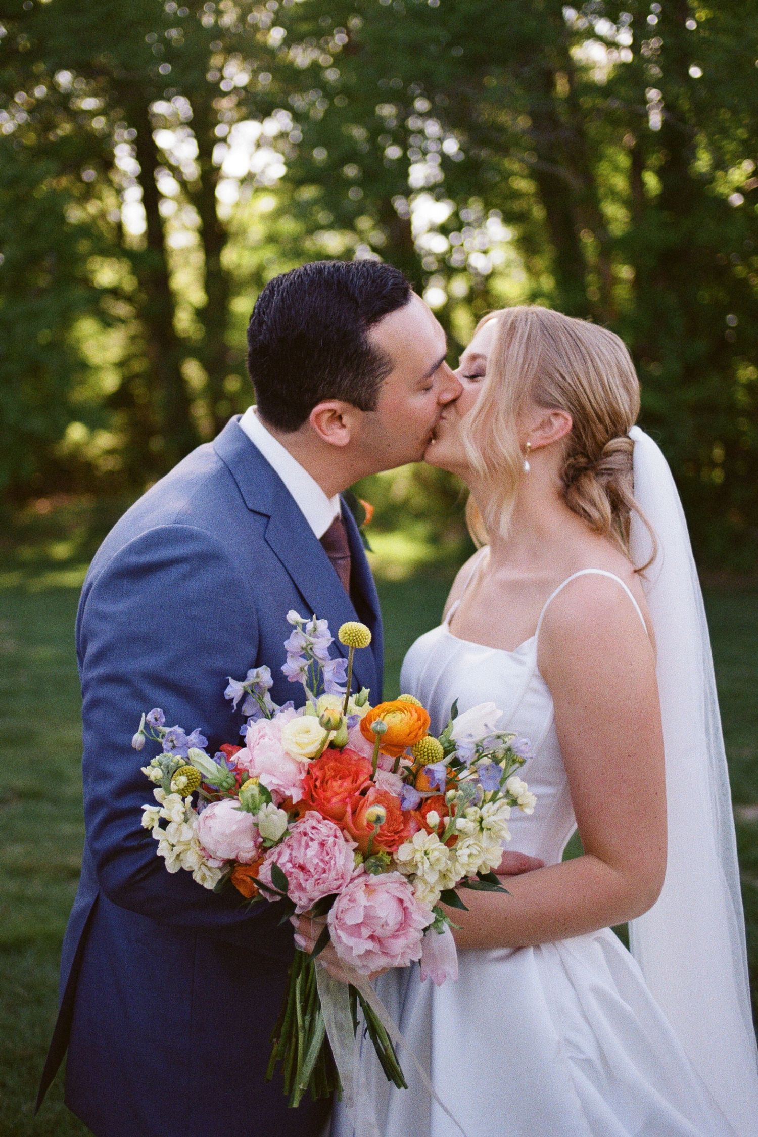 Bride and groom share a kiss outdoors, bride holds colorful bouquet with peonies and ranunculus, groom wears blue suit.