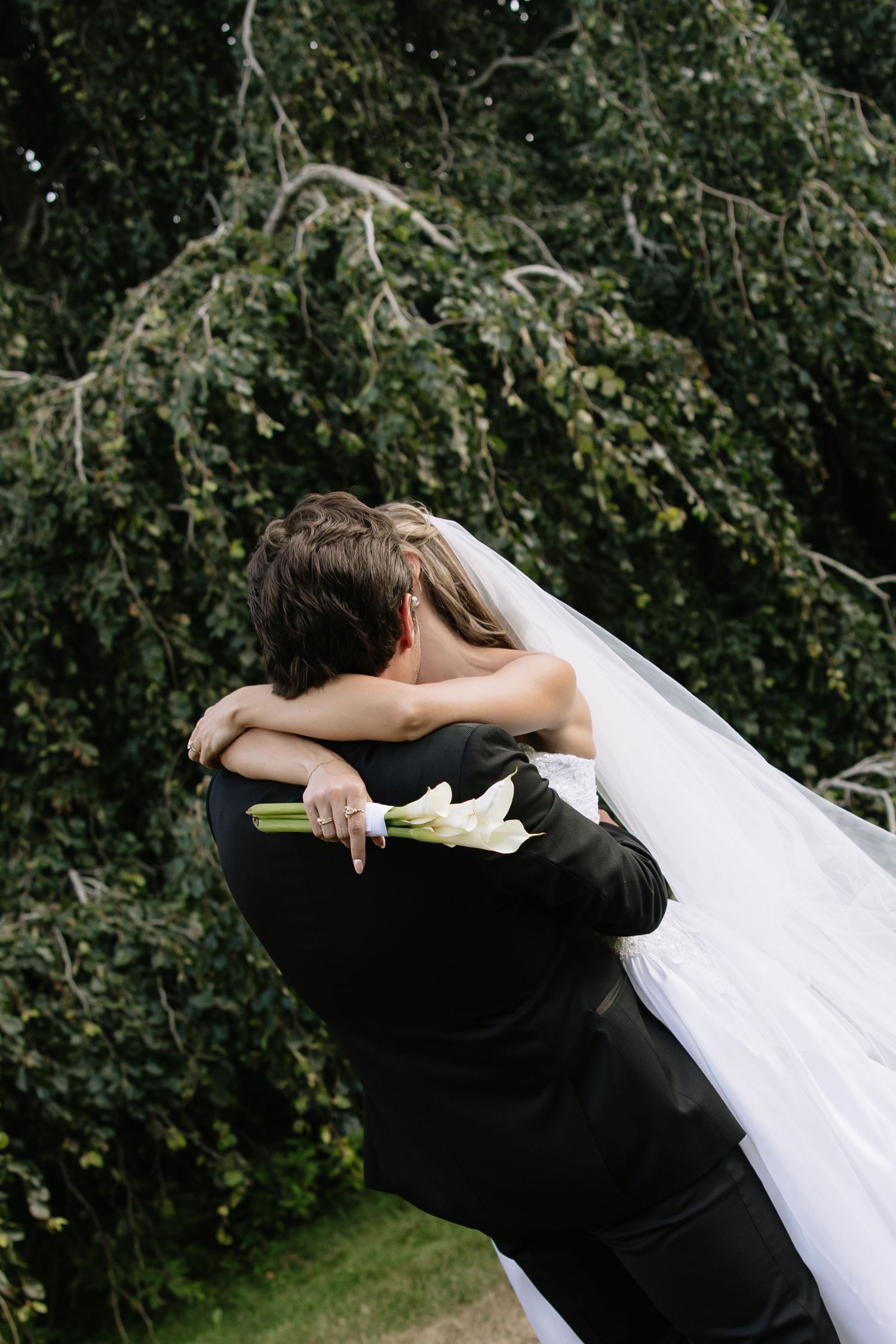 Bride and groom embracing outdoors, bride holding white calla lilies bouquet with veil flowing, lush green trees in background.