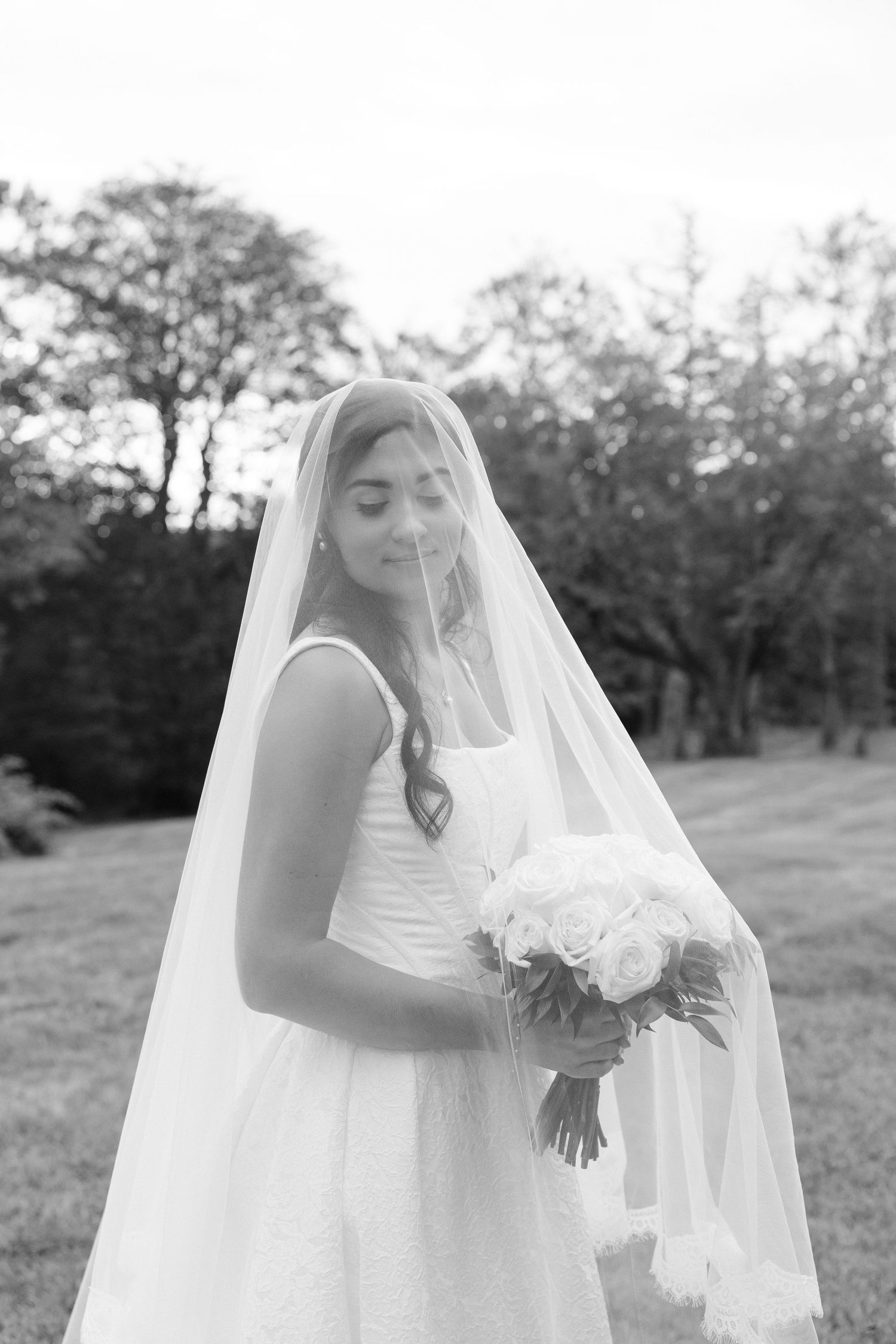 Bride in white gown with long veil holding bouquet of white roses, standing outdoors in black and white photo.