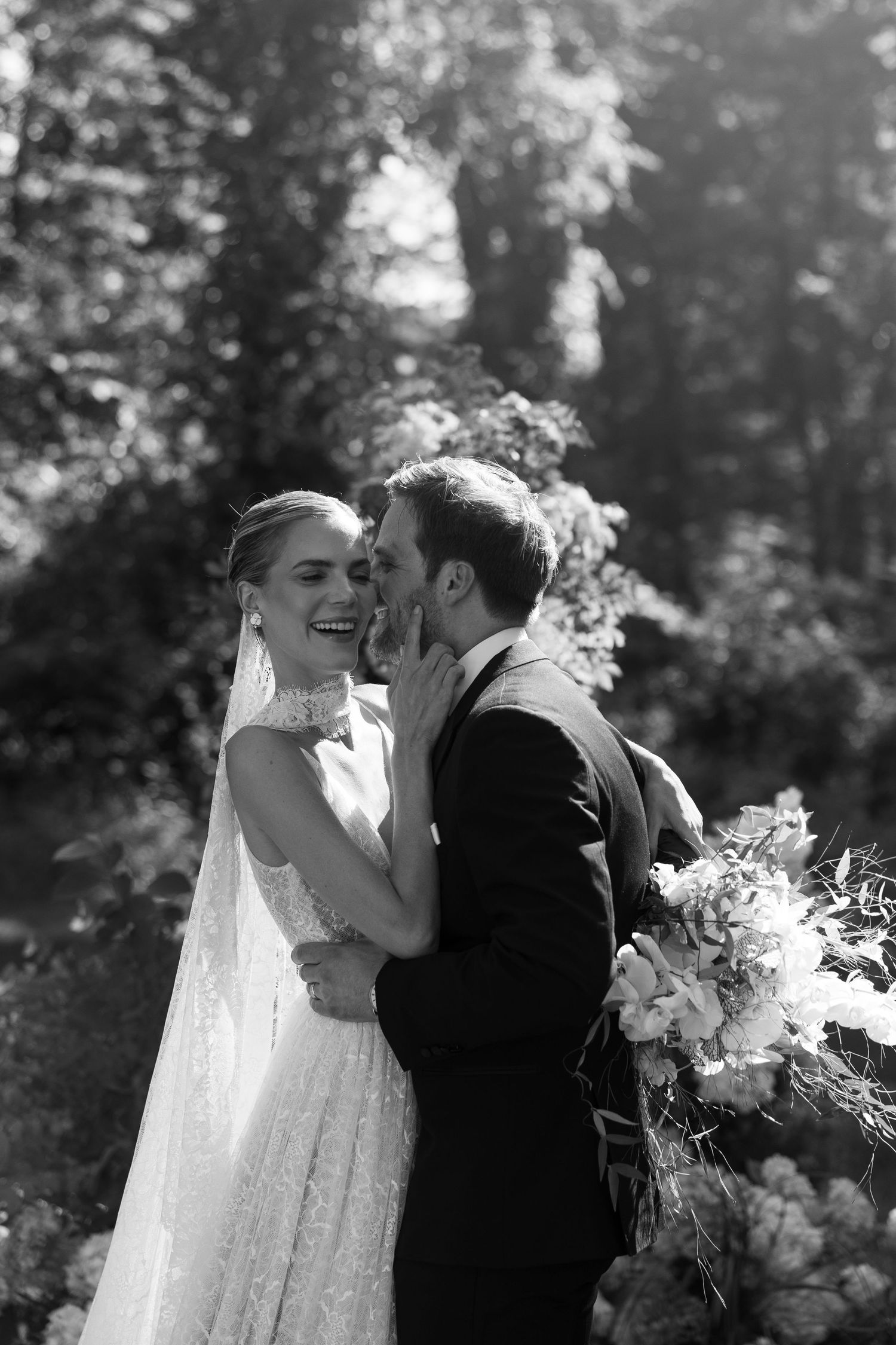 Black and white photo of a groom whispering to a laughing bride holding a bouquet in a lush garden.