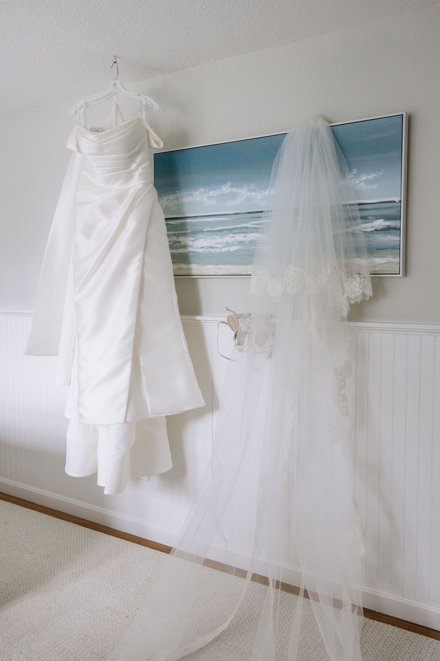 White wedding dress and lace veil hanging in a bright room with an ocean painting on the wall.