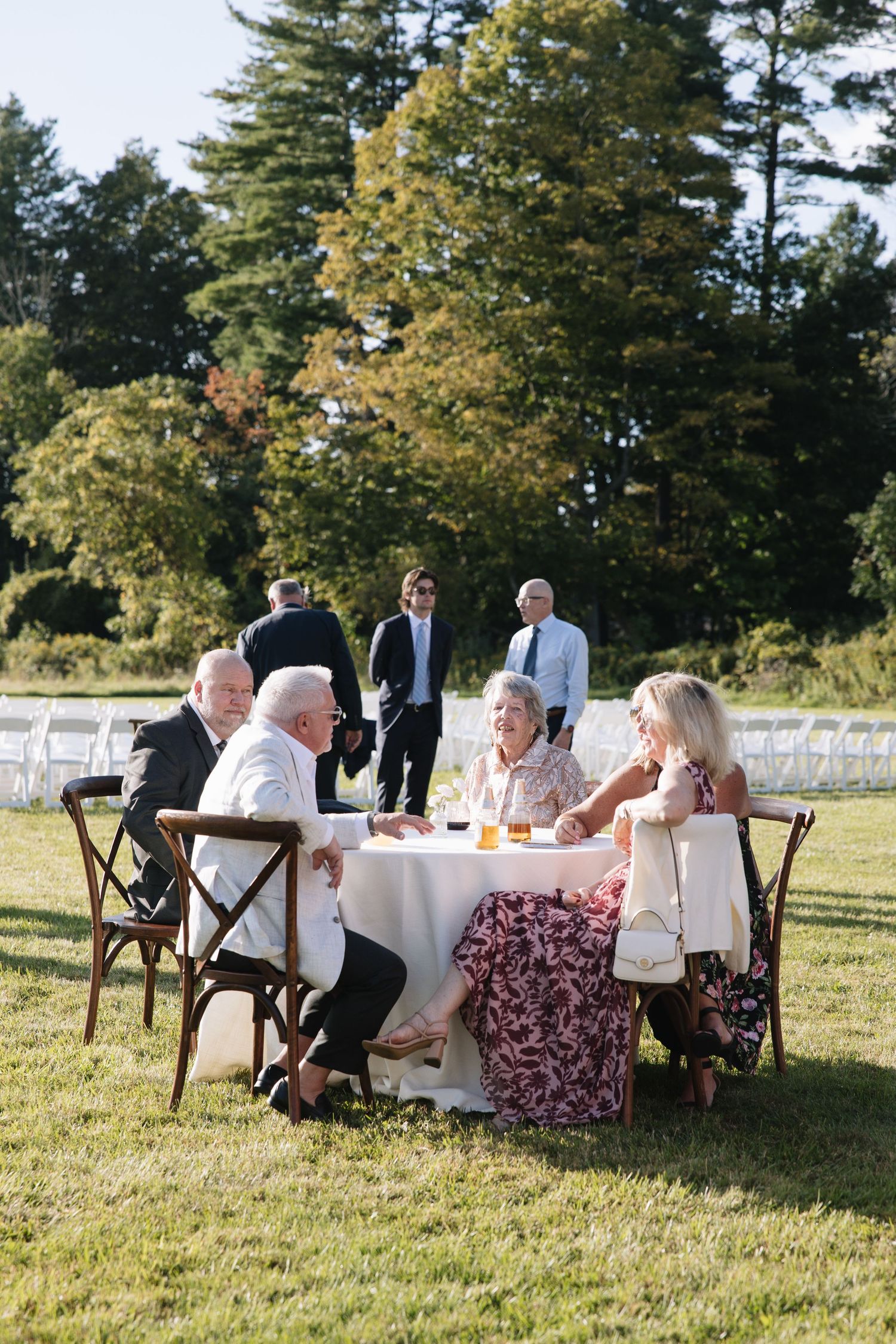 Elderly guests seated at outdoor wedding reception table on grass, with trees and pond in background.
