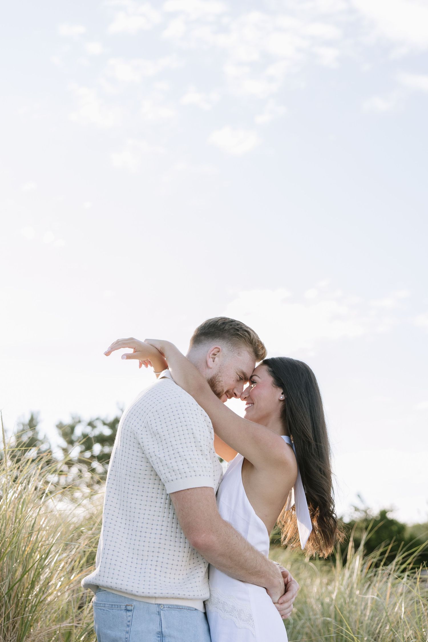 Romantic couple embracing in golden grass field, touching foreheads and smiling under a bright sky.