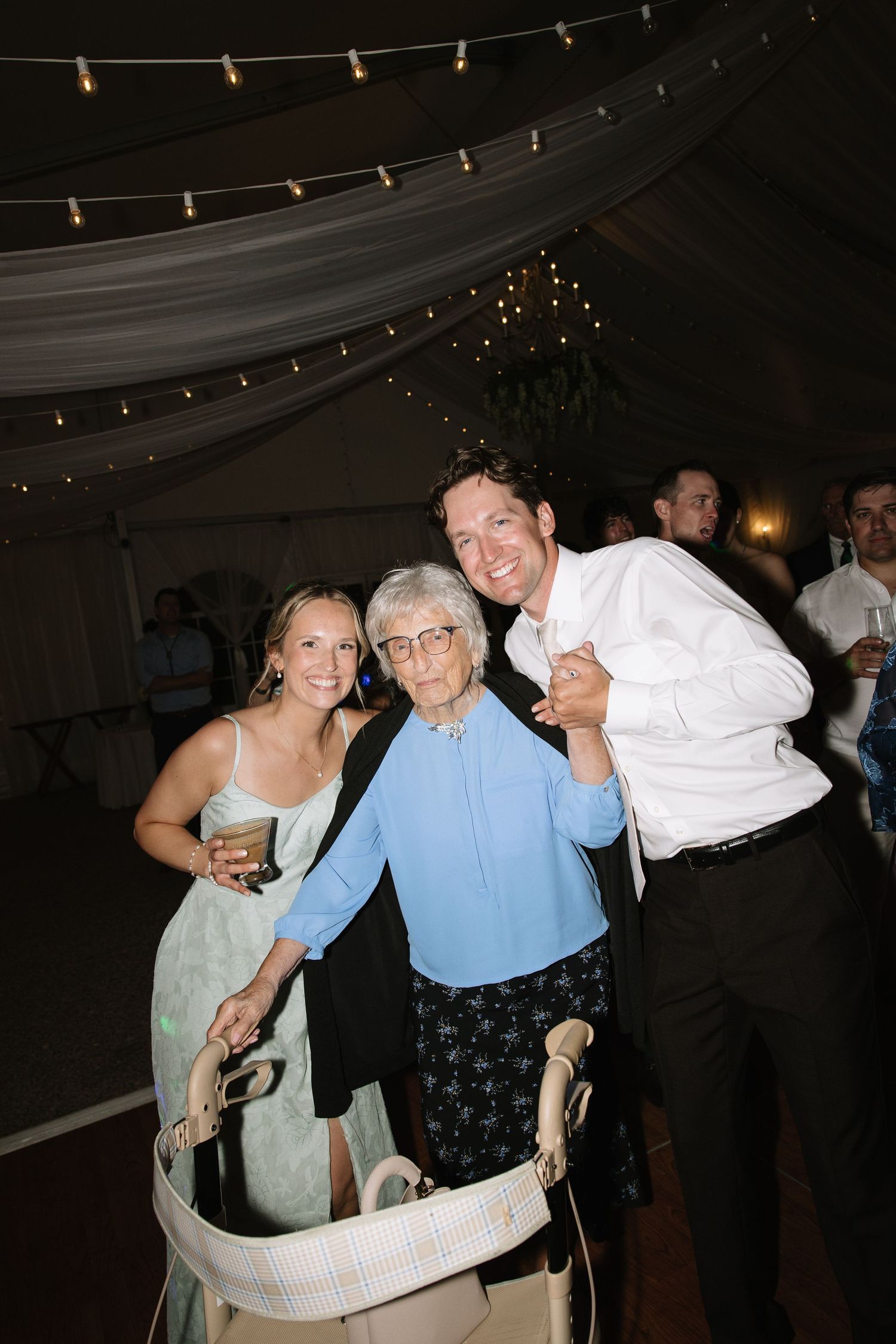 Elderly woman with walker poses with young couple at a festive wedding reception under string lights.