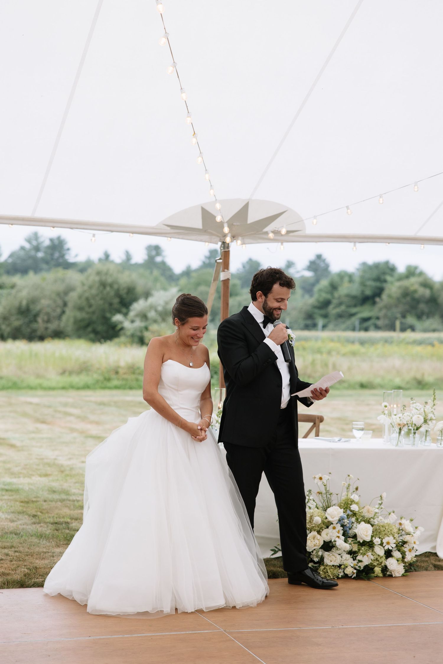 Bride smiles as groom reads heartfelt vows under a white tent at outdoor wedding reception.