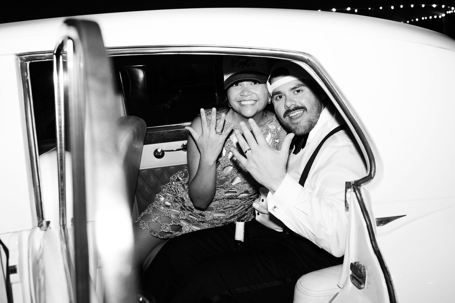 Black and white photo of a couple in a limo showing off rings, celebrating their engagement at night.