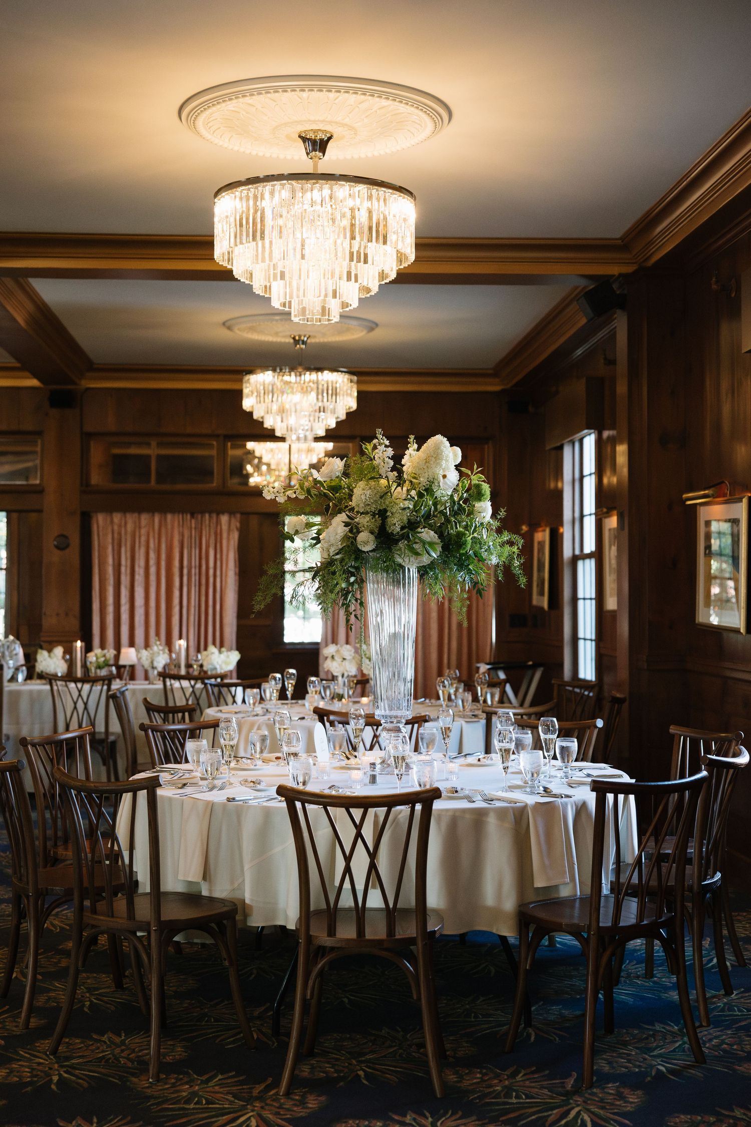 Elegant wedding reception room with crystal chandeliers, round tables, white linens, and tall floral centerpieces.