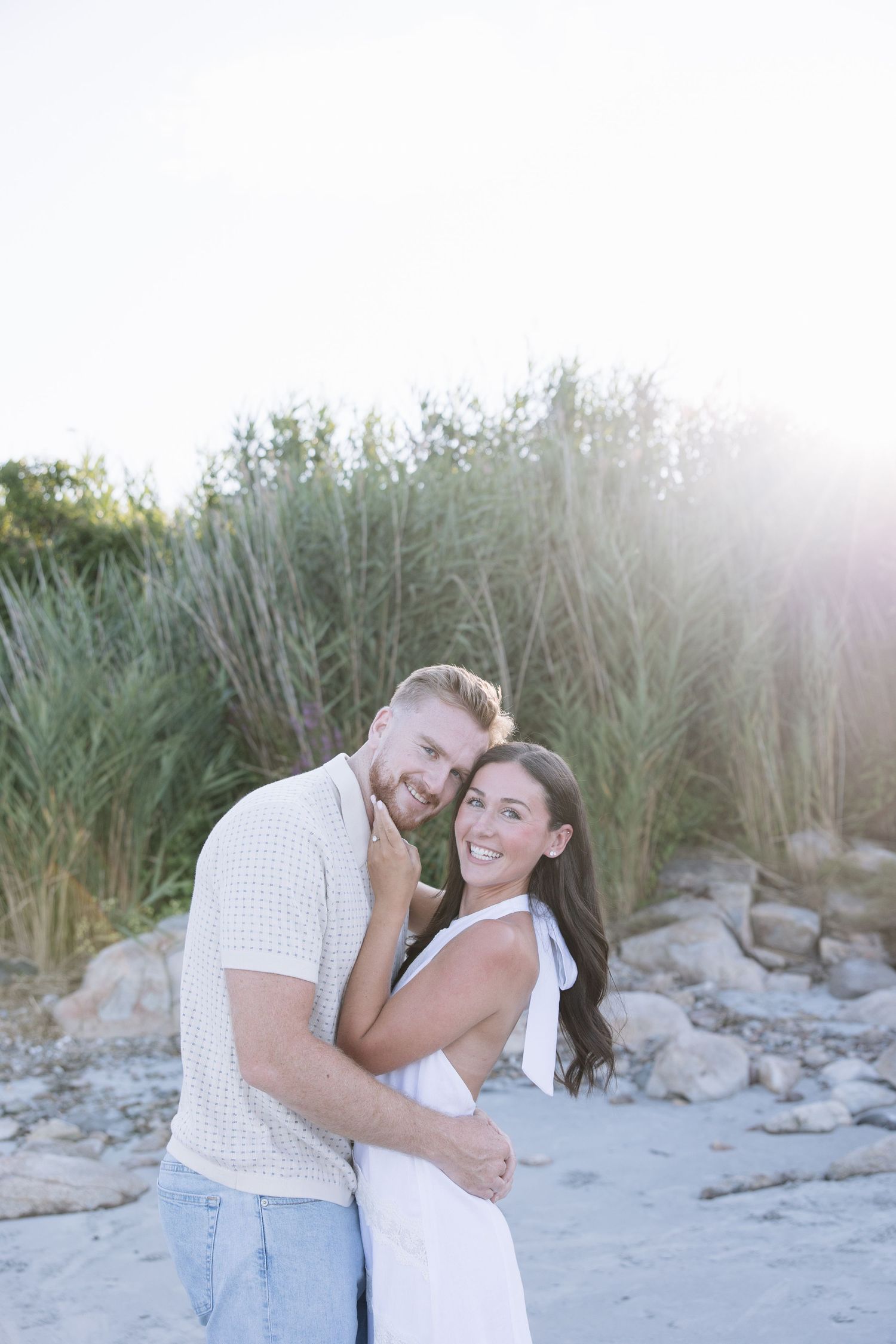 A smiling couple embraces on a sandy beach with tall green grass and warm sunlight behind them.