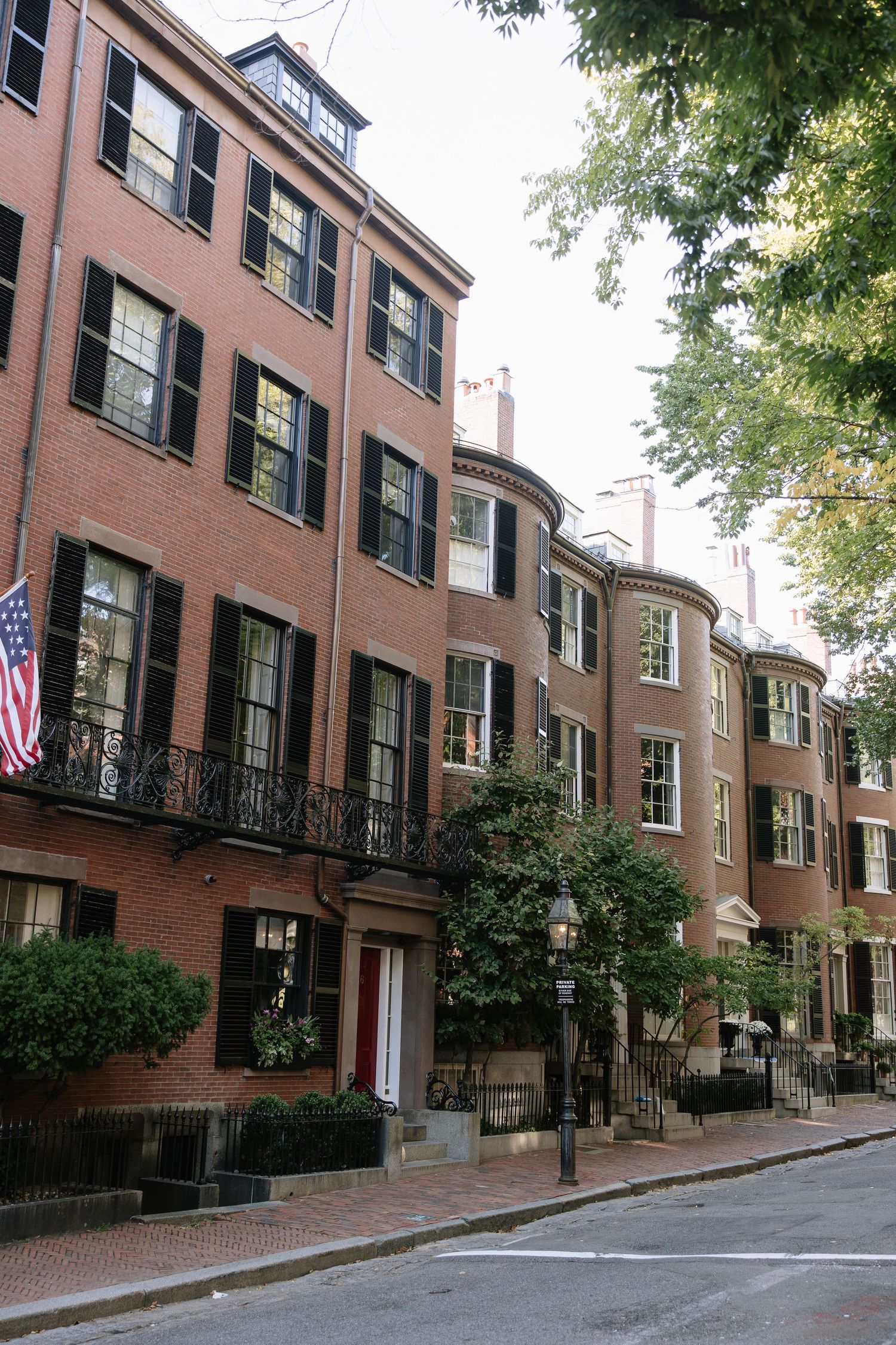 Historic brick townhouses with black shutters and American flag on a tree-lined Boston street.