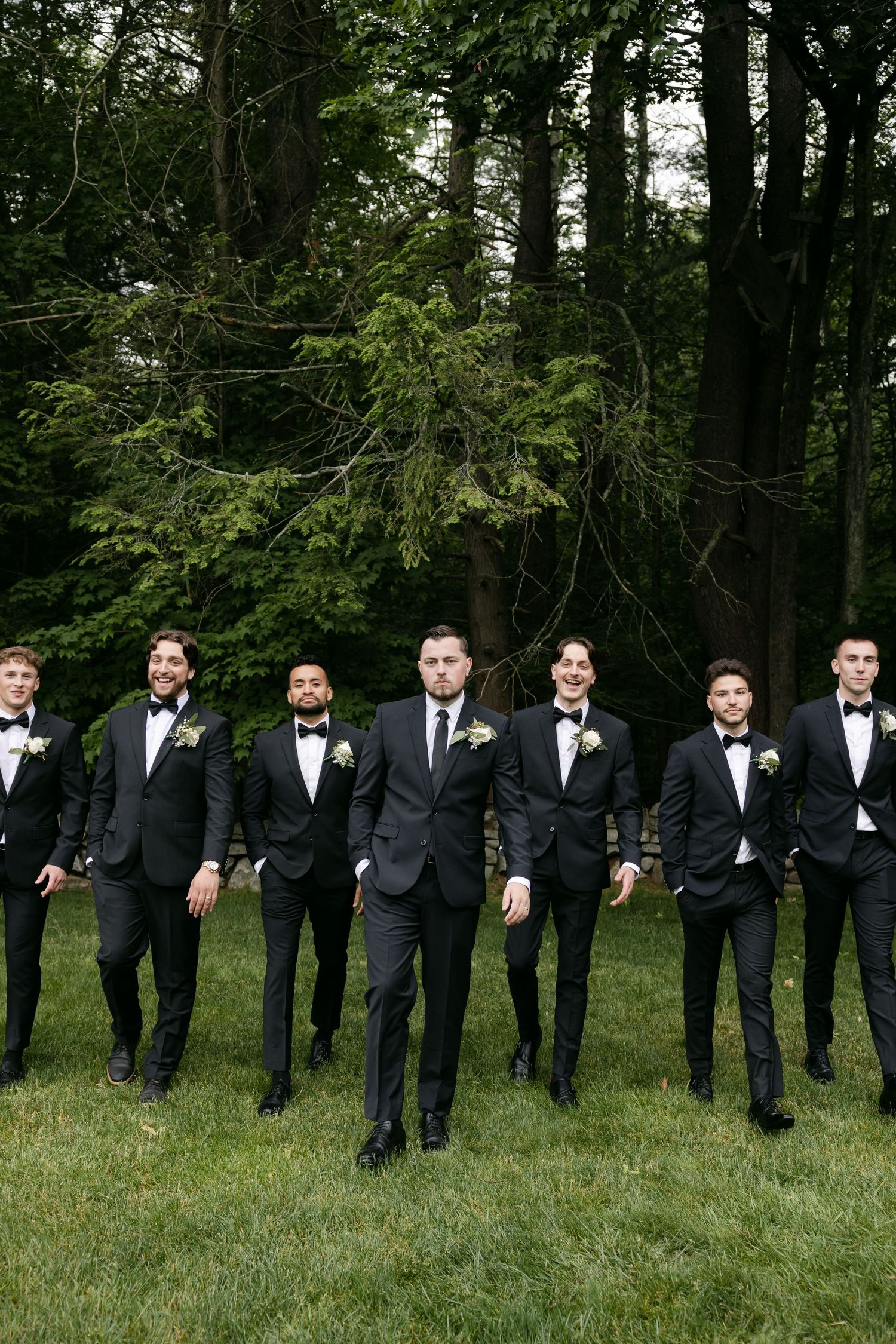 Row of groomsmen in black tuxedos stand together on grass against wooded backdrop.