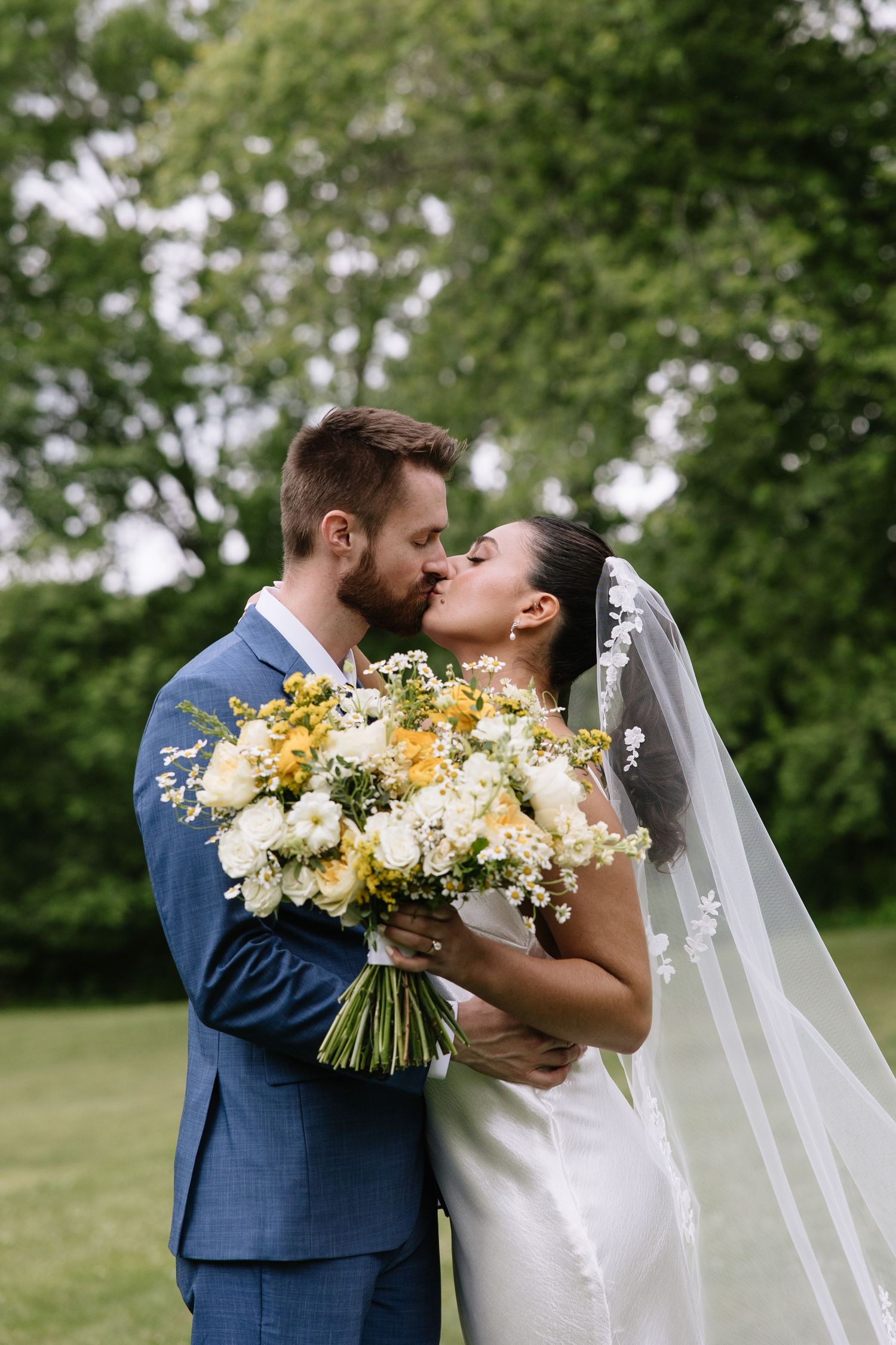 Bride and groom kissing outdoors, bride holding yellow and white floral bouquet, groom in blue suit.