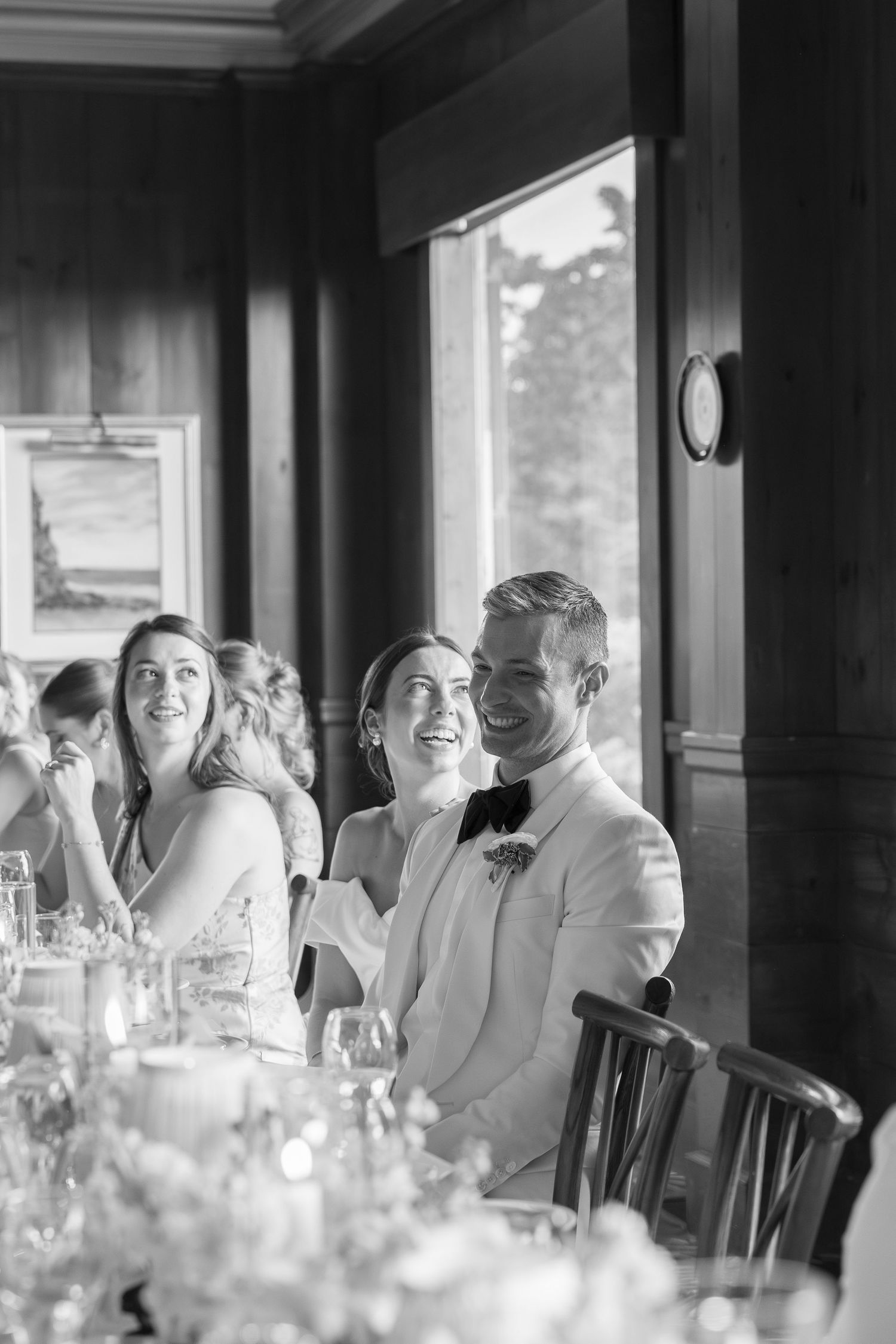 Black and white wedding reception photo of smiling bride and groom seated at head table with guests.