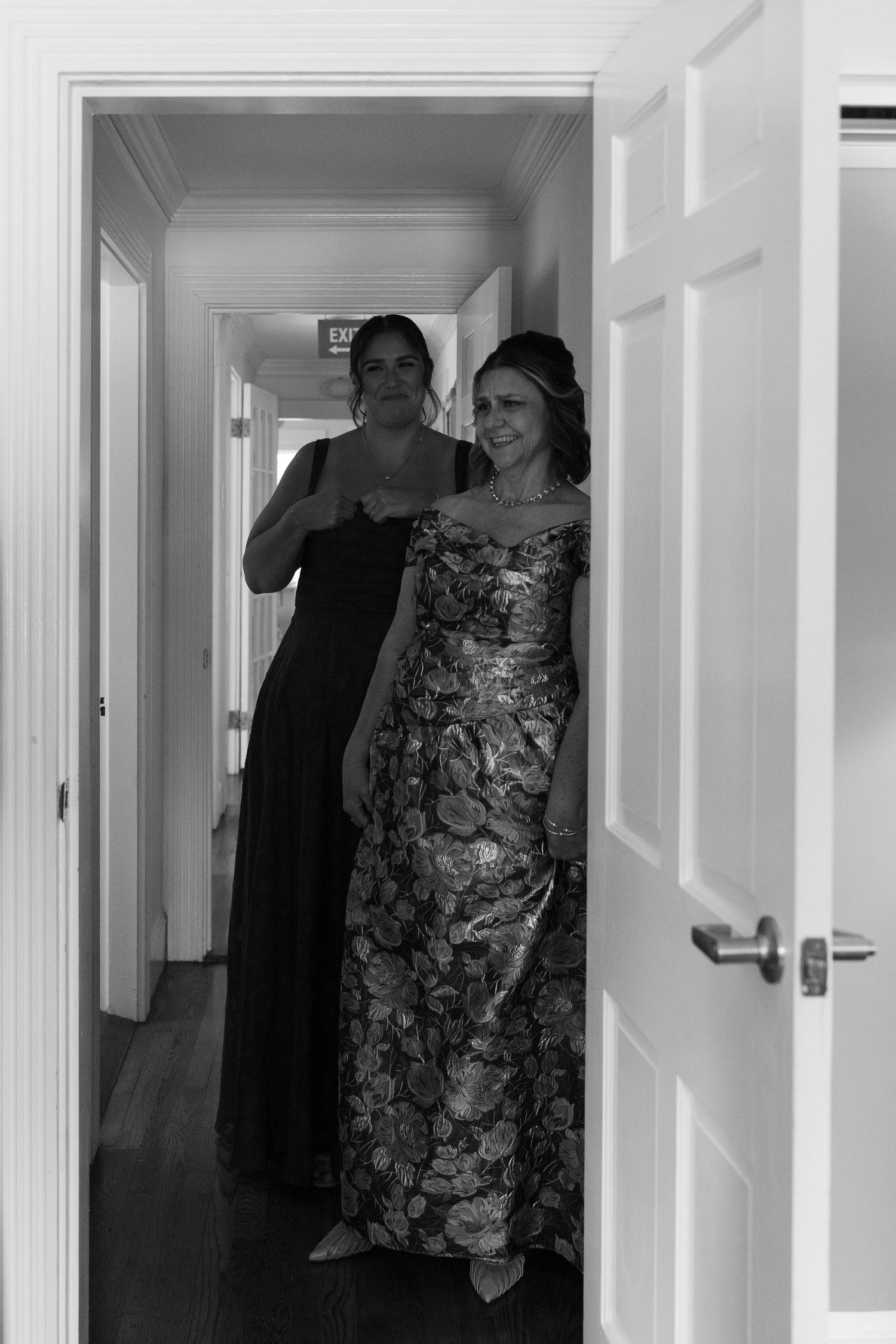 Two elegant women in formal gowns pose in a hallway, captured in black and white photography.