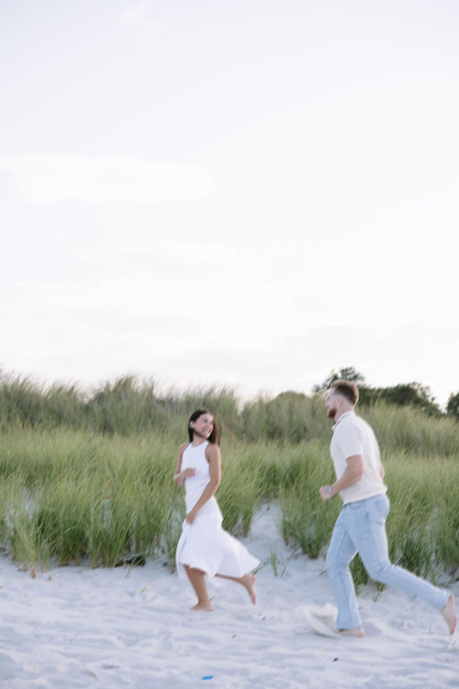 A couple runs barefoot on a sandy beach, the woman in a white dress laughing as they play near green dunes.