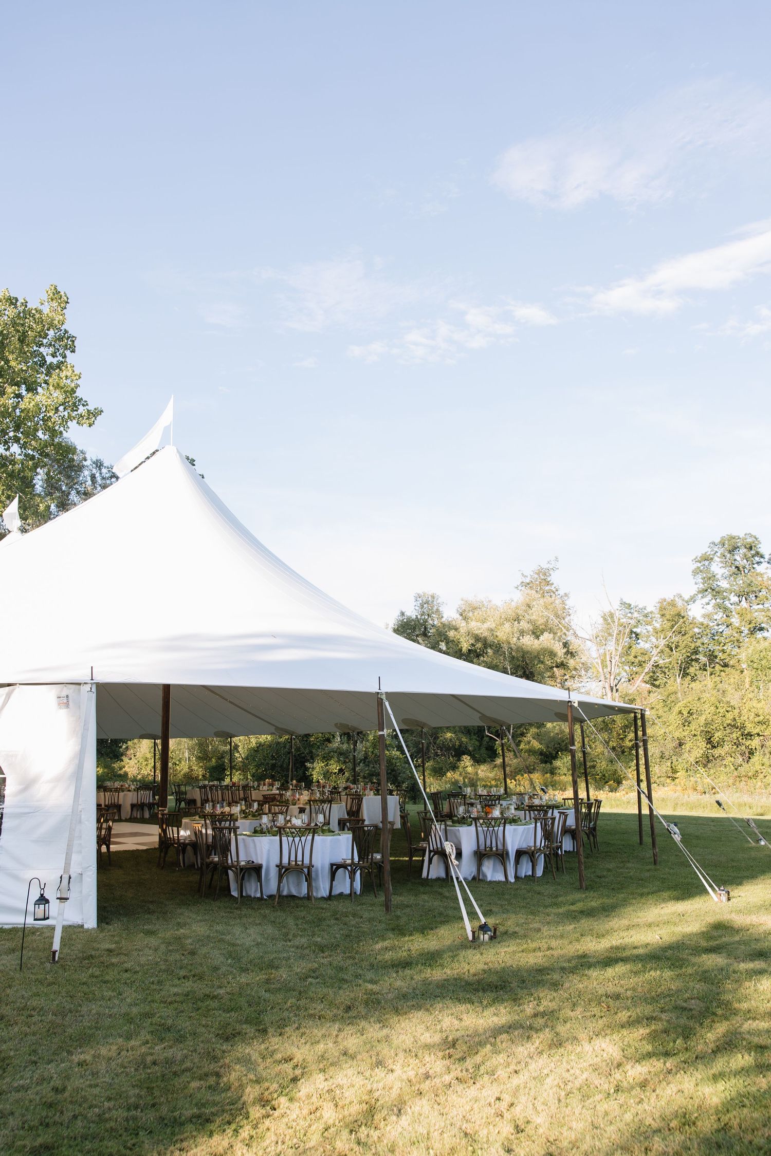 White high-peak wedding reception tent set on lush green lawn with round tables and chairs arranged underneath on a sunny day.