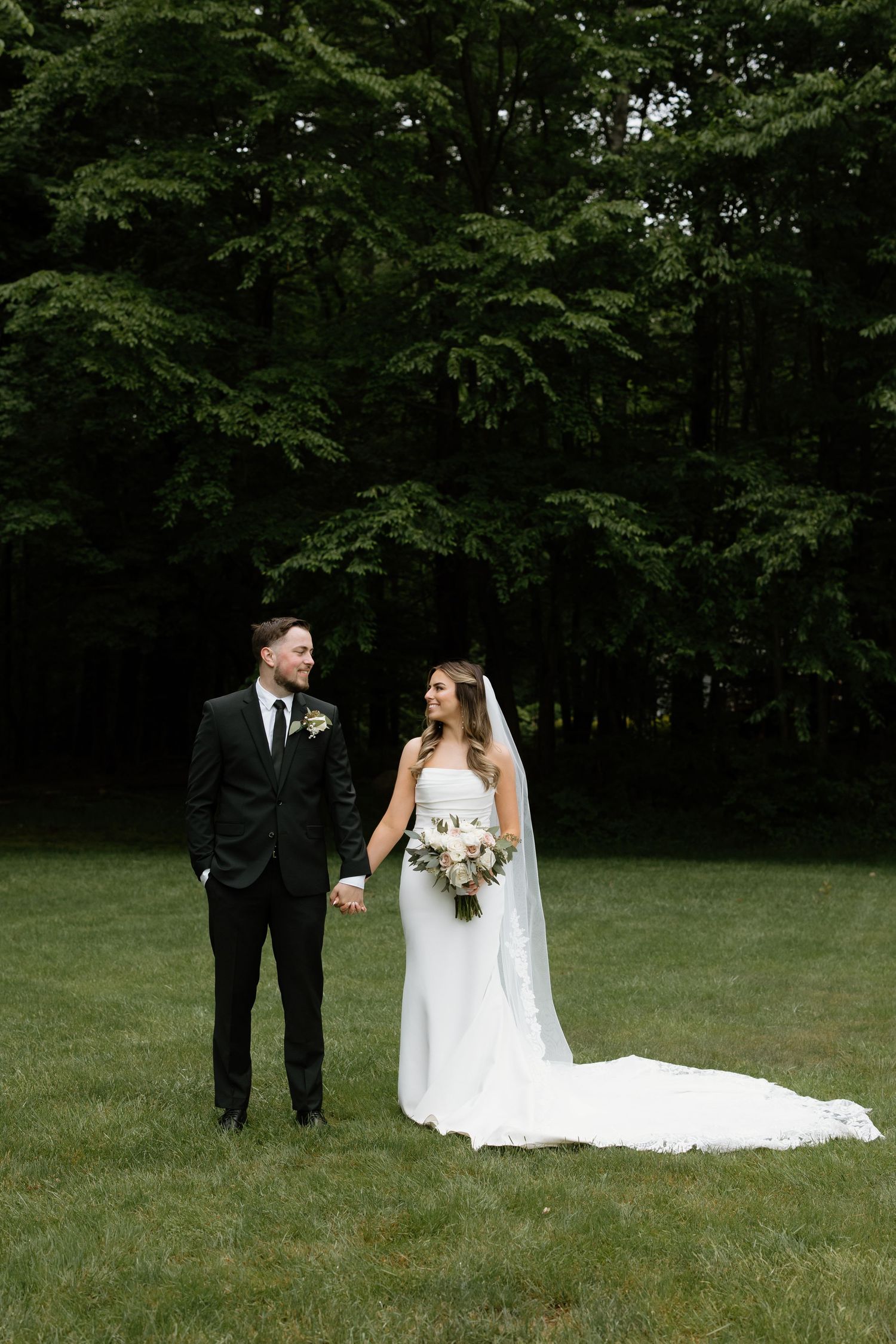 Wedding couple poses in grass field with bride holding bouquet and wearing flowing white gown.