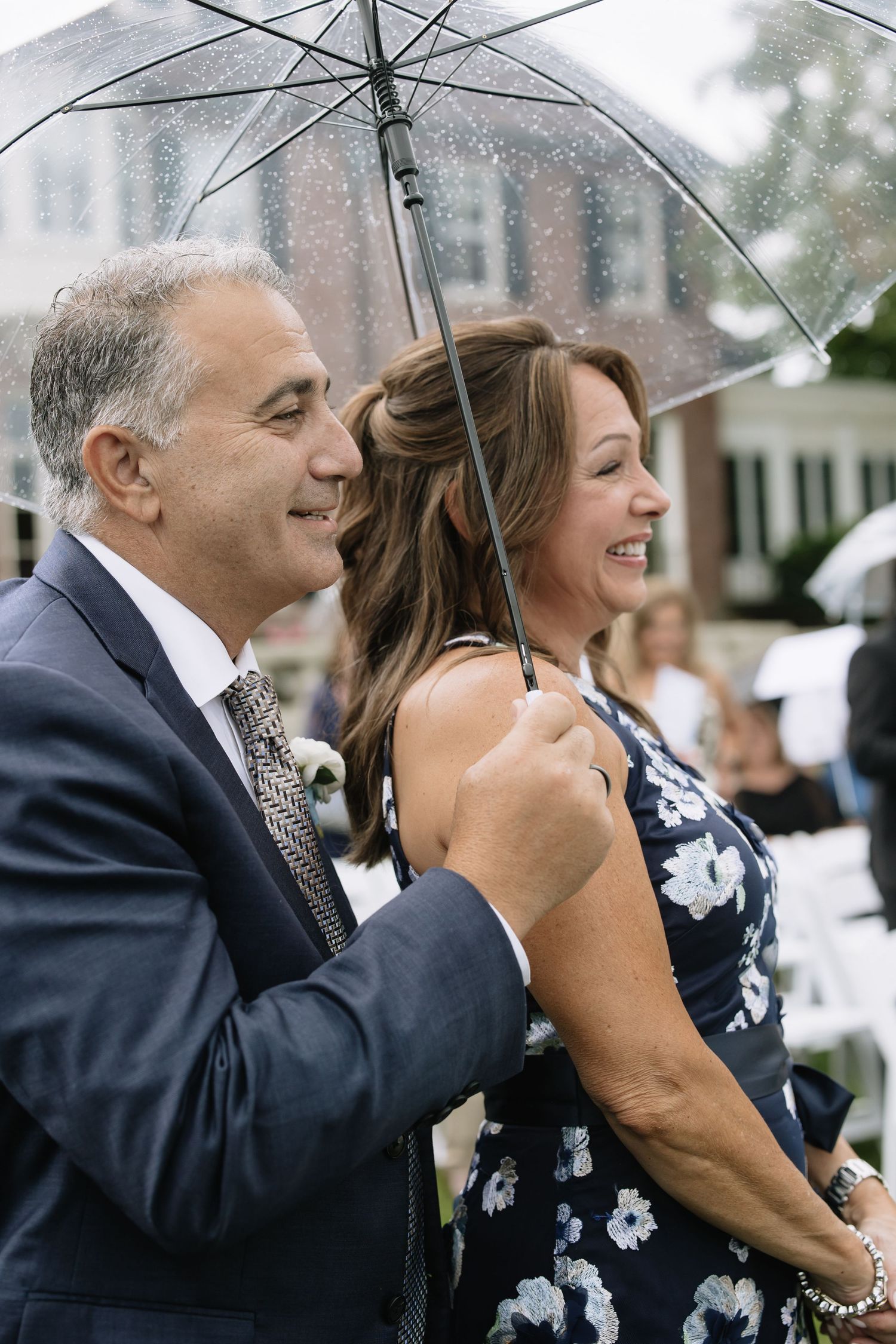 Wedding guests share a clear umbrella while watching the ceremony in the rain.