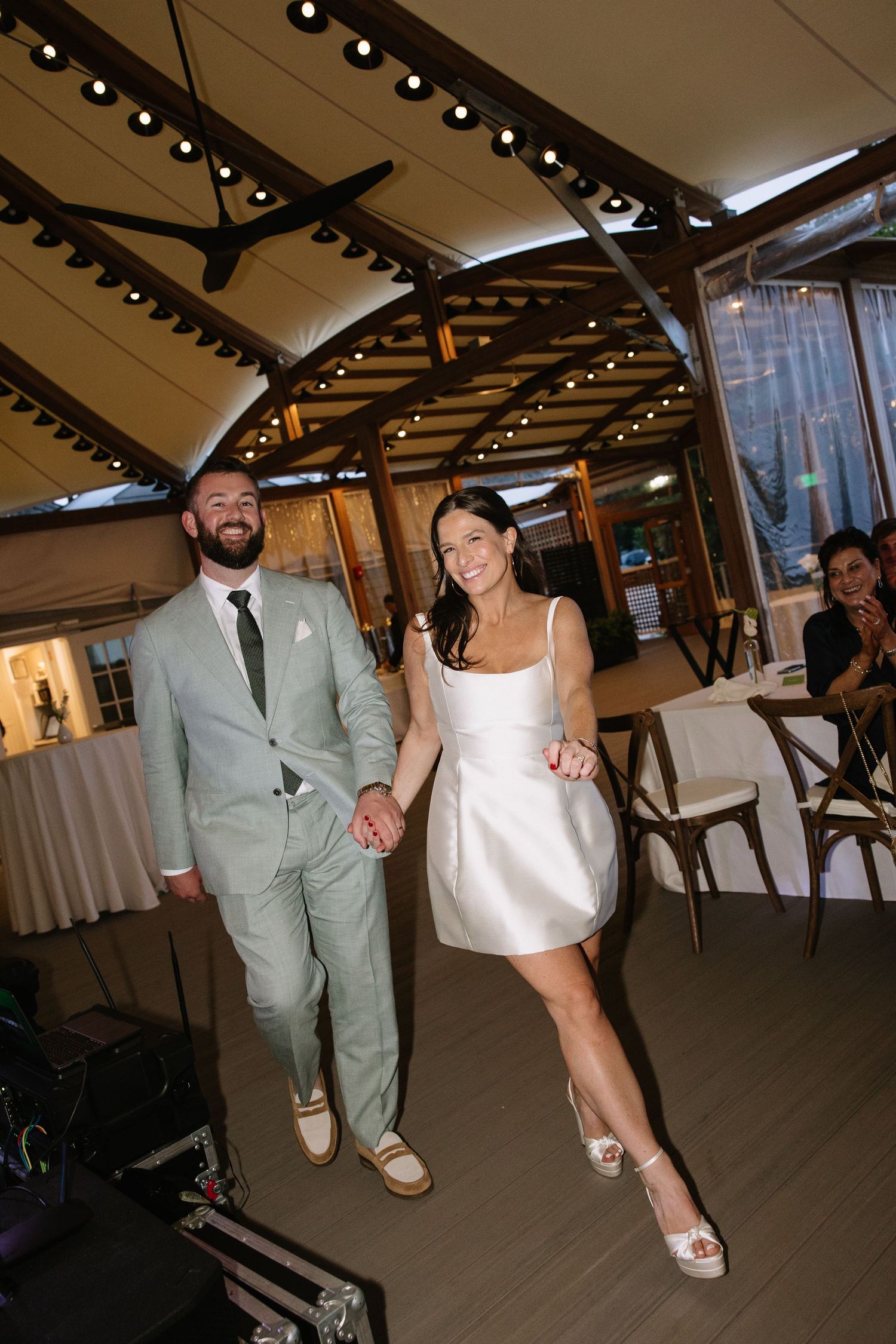 Bride in short white dress and groom in sage green suit hold hands walking into wedding reception venue.