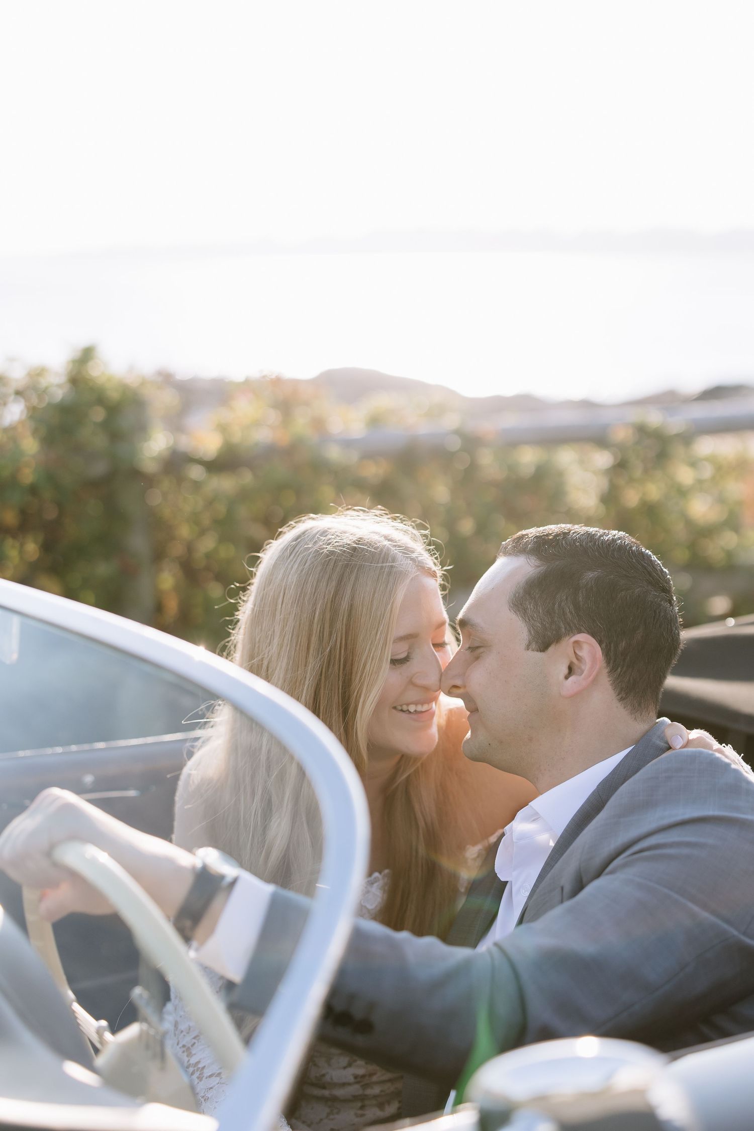 Couple sharing a romantic moment in a vintage convertible car with scenic countryside views in warm sunlight.