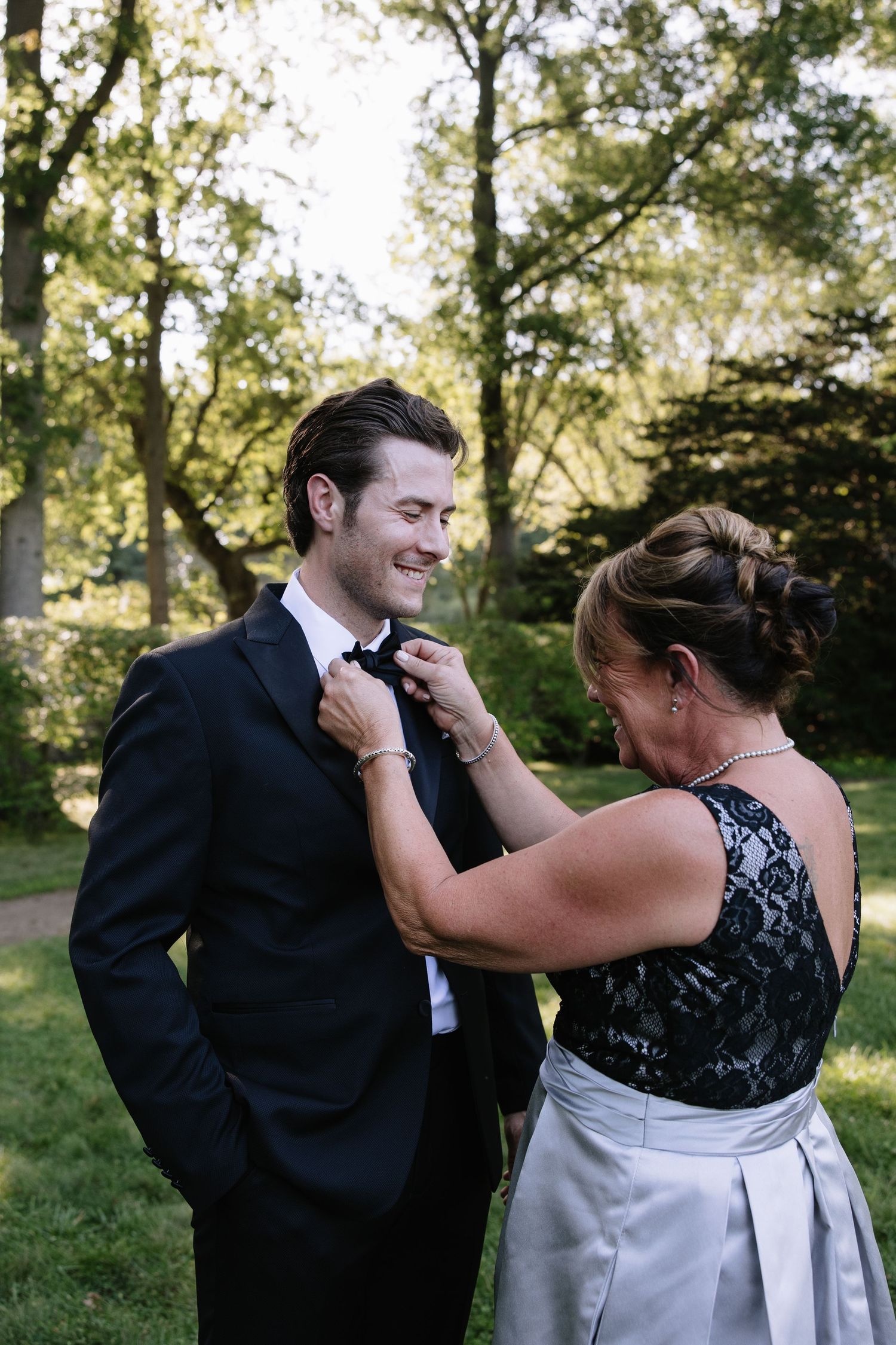 A woman in black lace adjusts a groom's bow tie outdoors, surrounded by lush green trees.