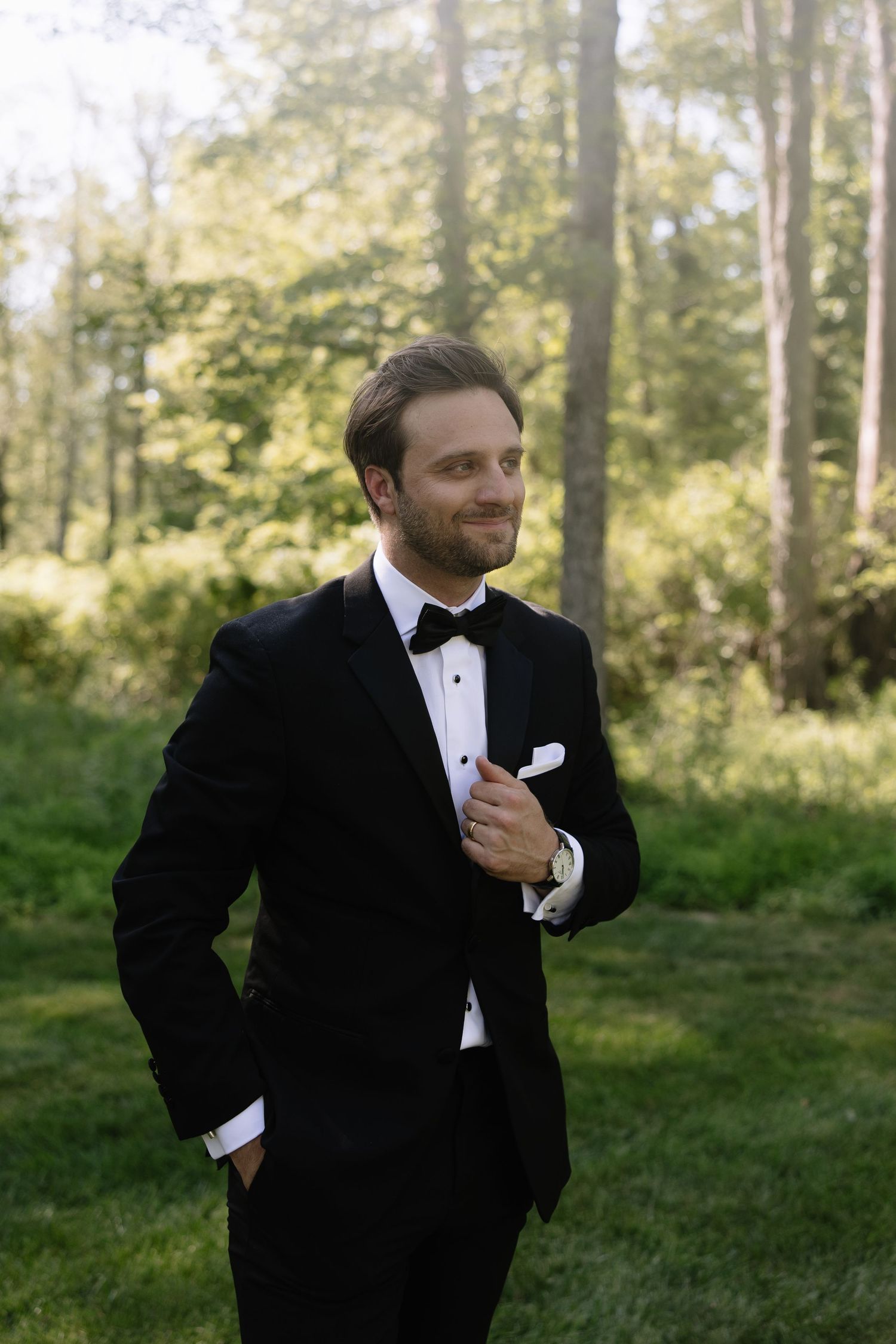 Handsome groom in black tuxedo smiling outdoors in a lush green forest setting.
