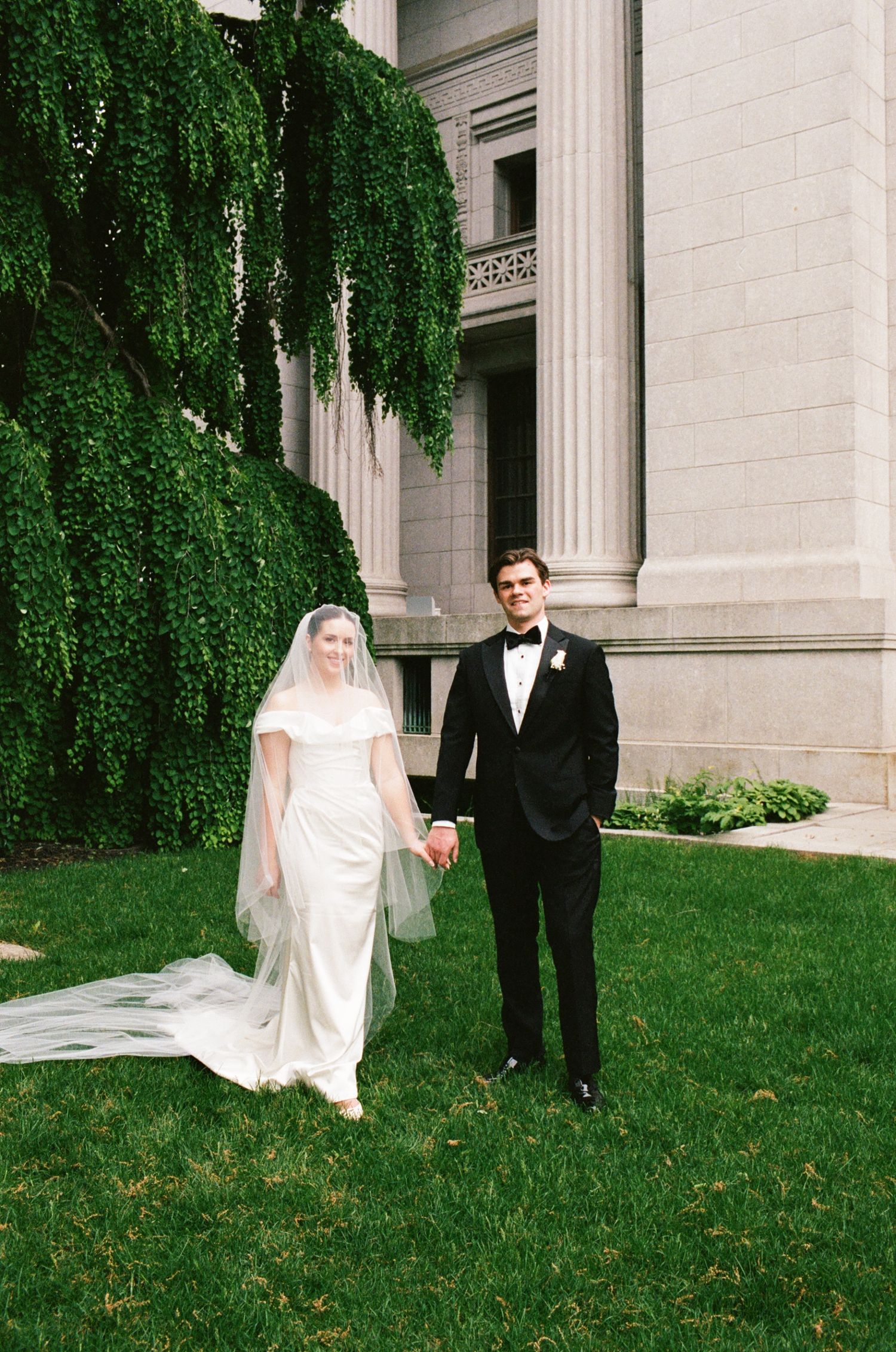 Bride in white gown with veil and groom in black tuxedo holding hands on green lawn beside stone building.